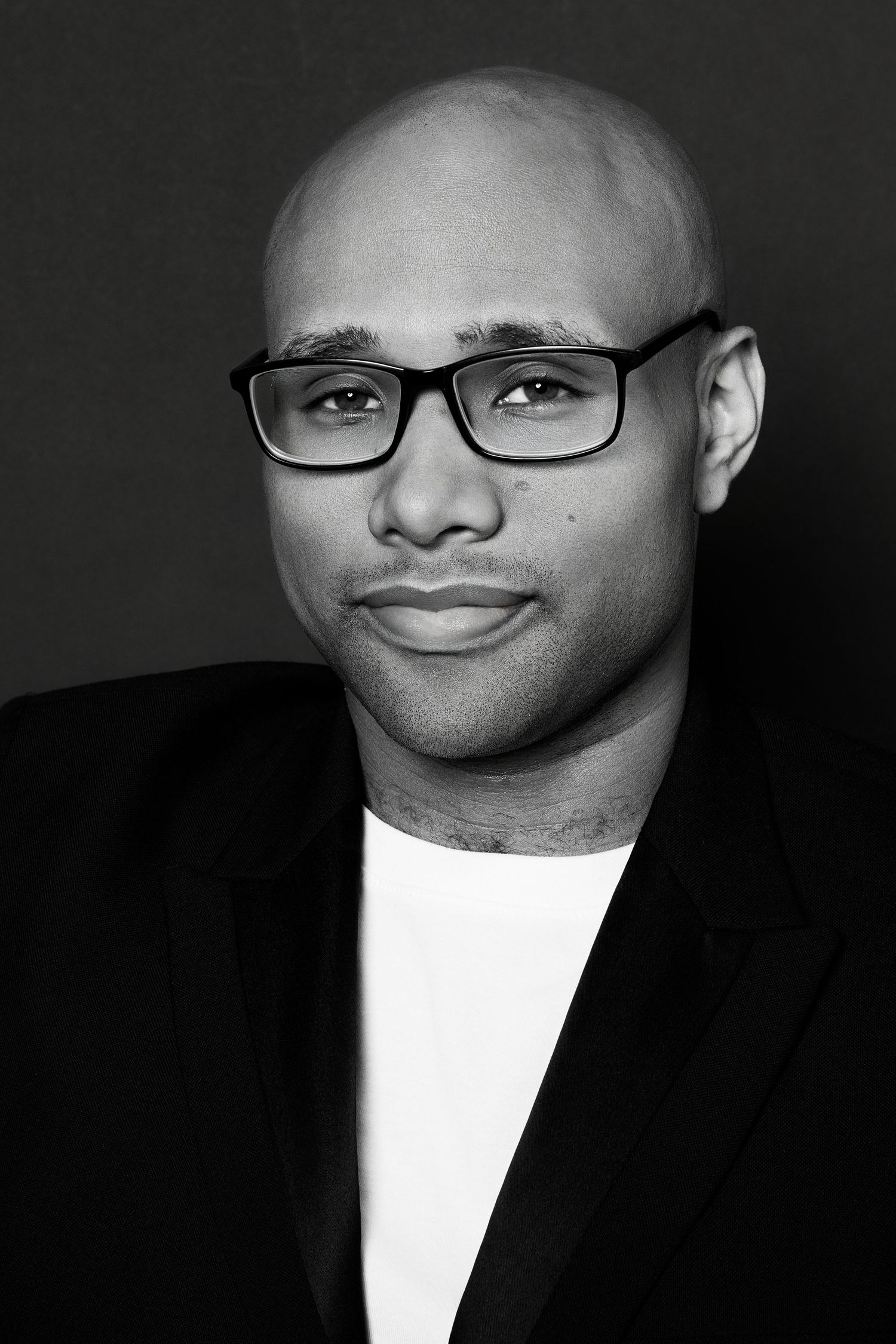 Bald Black man in black blazer wearing rectangular glasses with slight smile, black and white portrait photo.