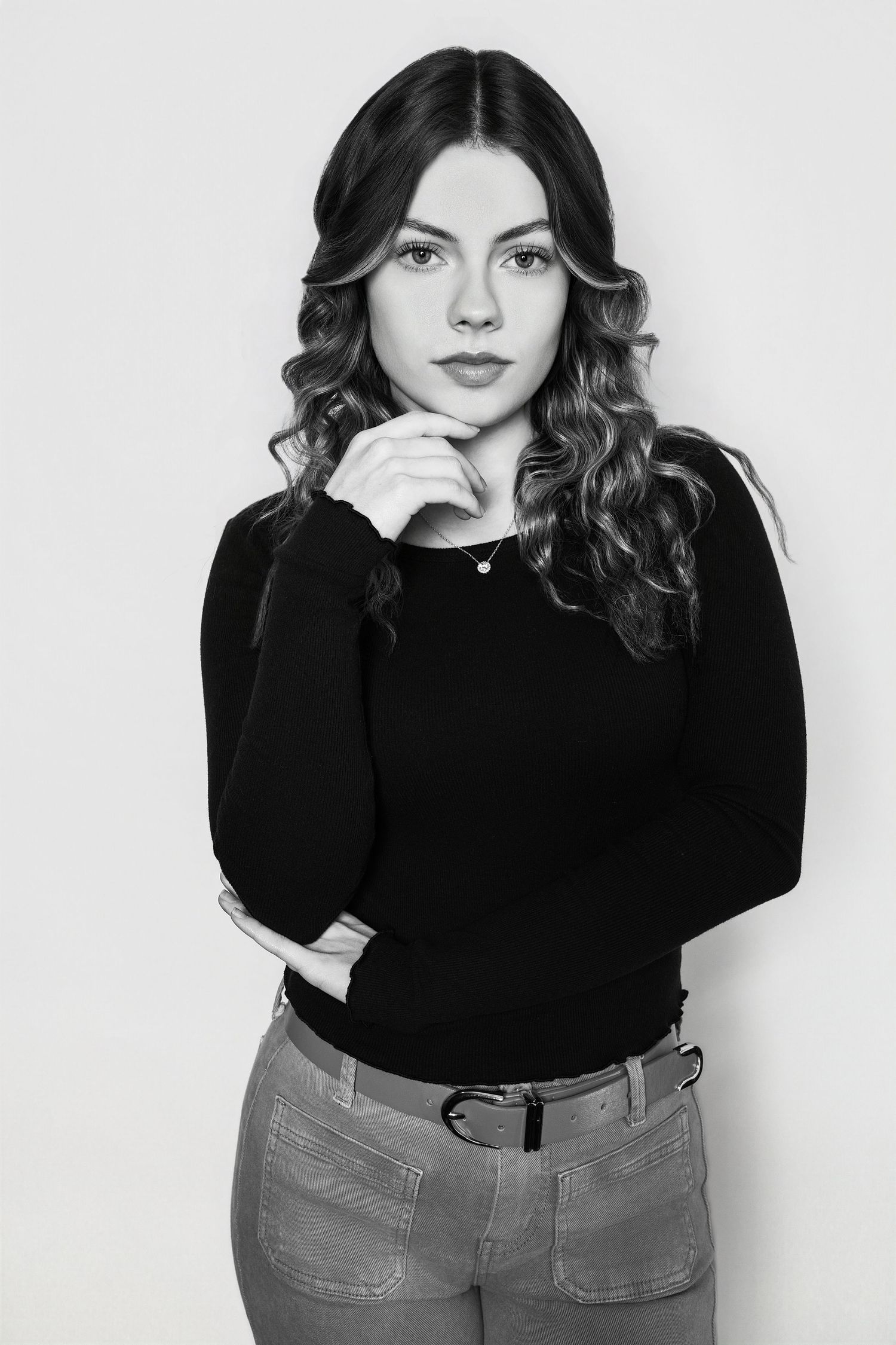 Black and white portrait of a young woman in black top and jeans posing thoughtfully with hand on chin.