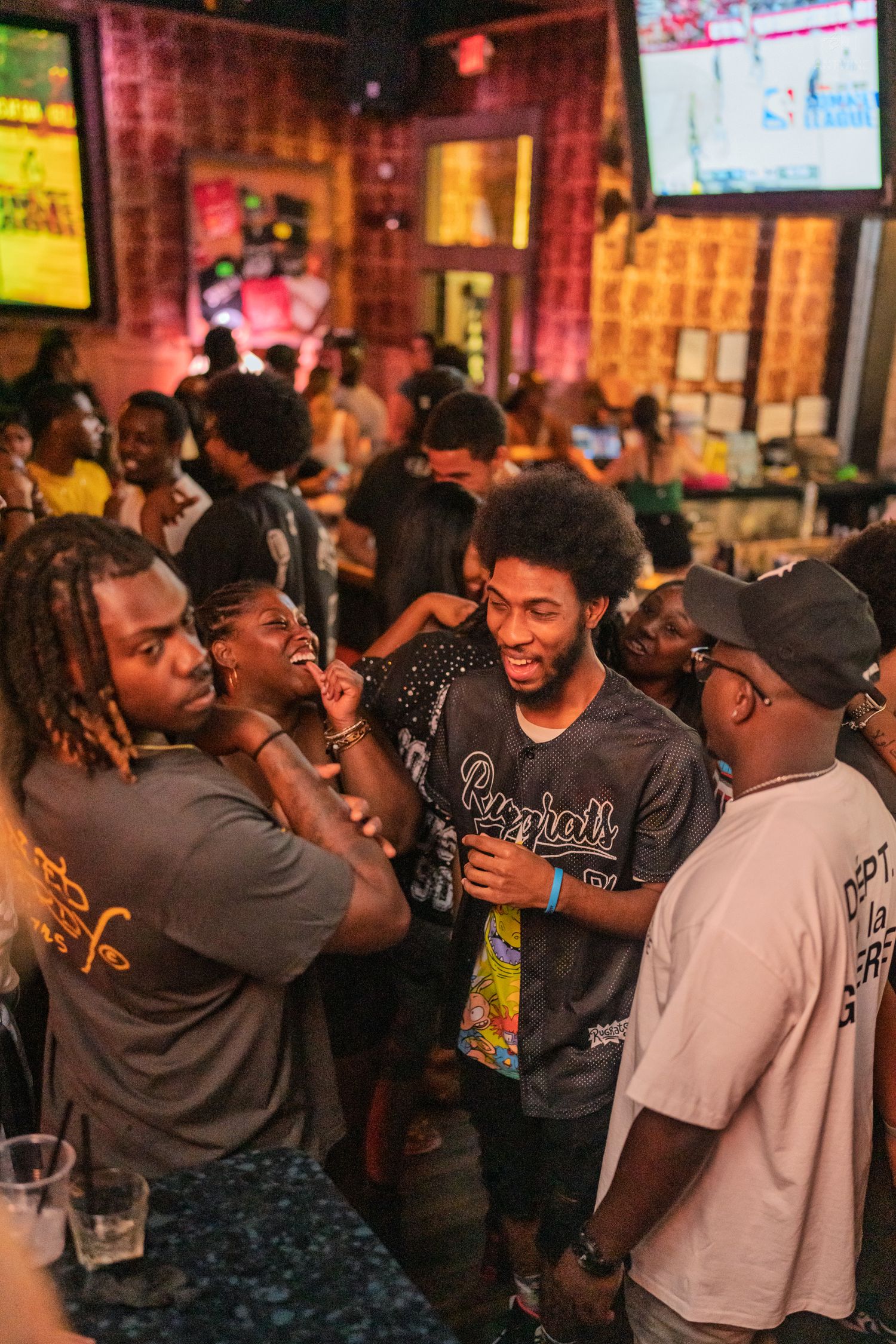 Group of young men laughing and socializing near a bar with sports TVs in background.