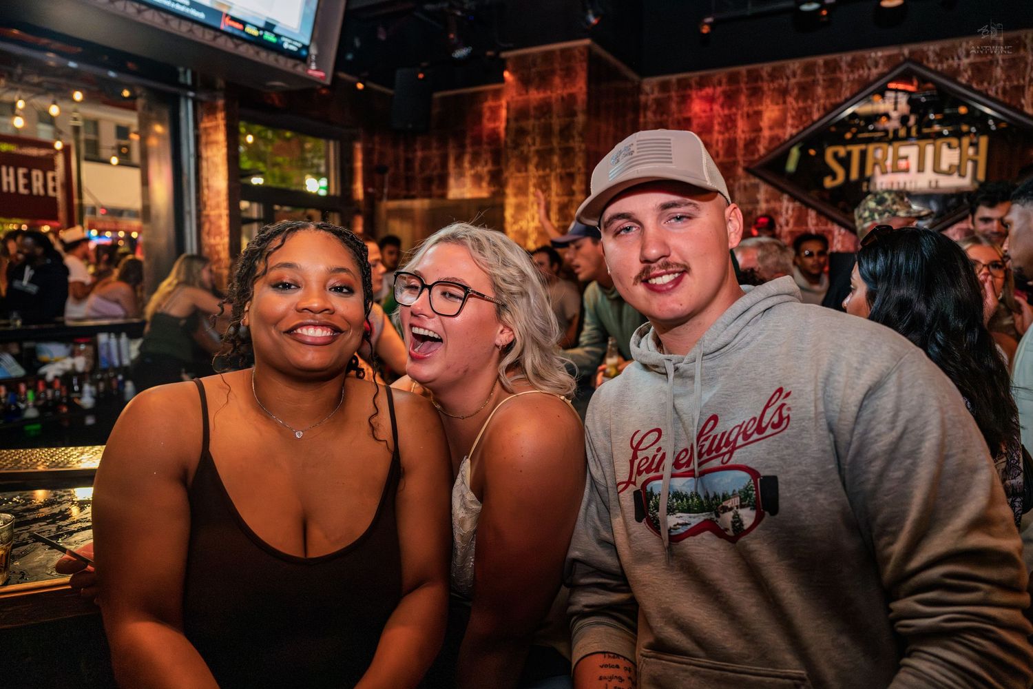 Three young adults smiling and posing together inside a lively bar with Stretch signage.