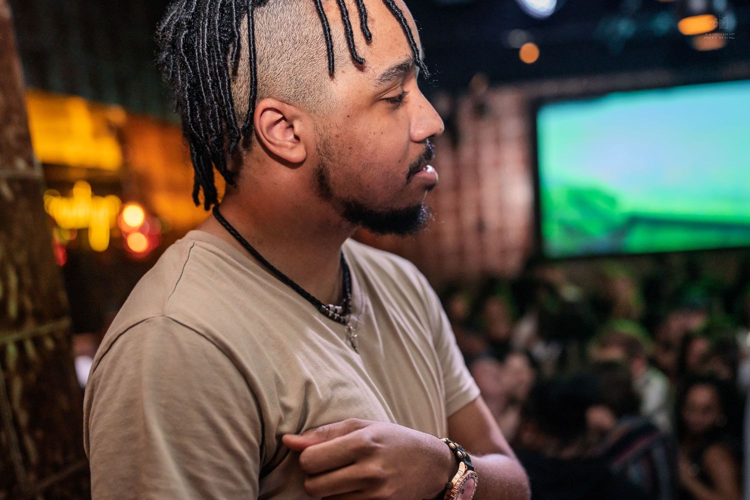 Side profile of a man with braids and a beaded necklace at a vibrant bar event.