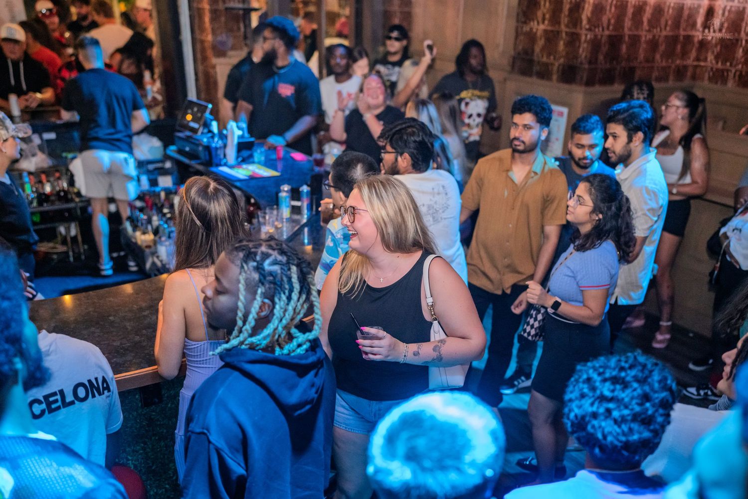 A diverse crowd mingles at a busy nightclub bar with blue lighting and a Barcelona jersey visible.