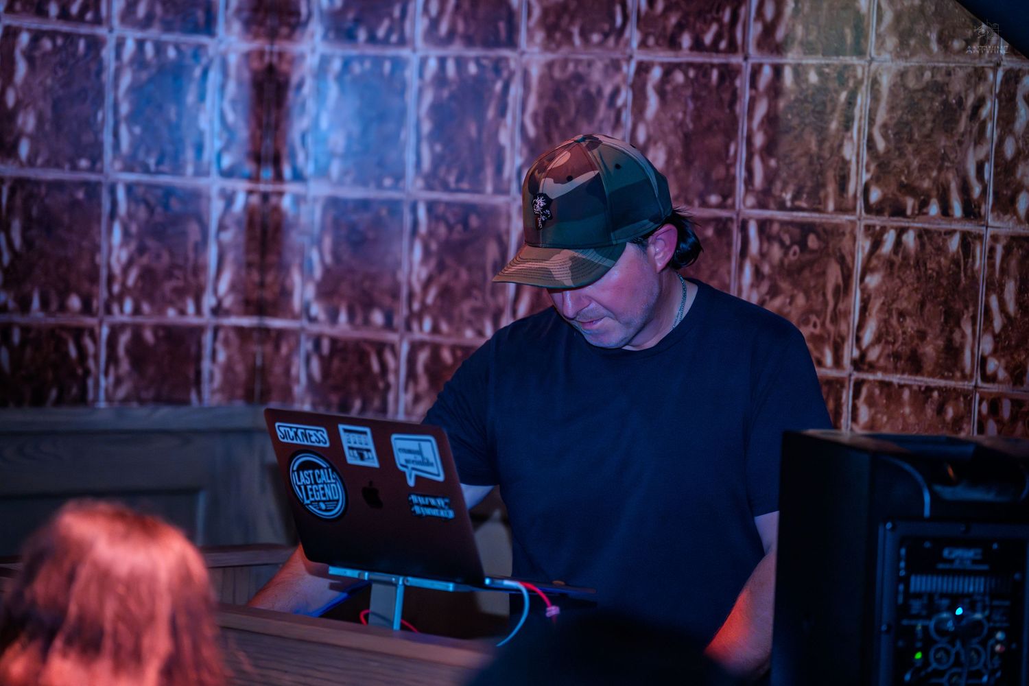 DJ working on a laptop with stickers at a nightclub, focused on mixing music in dim lighting.