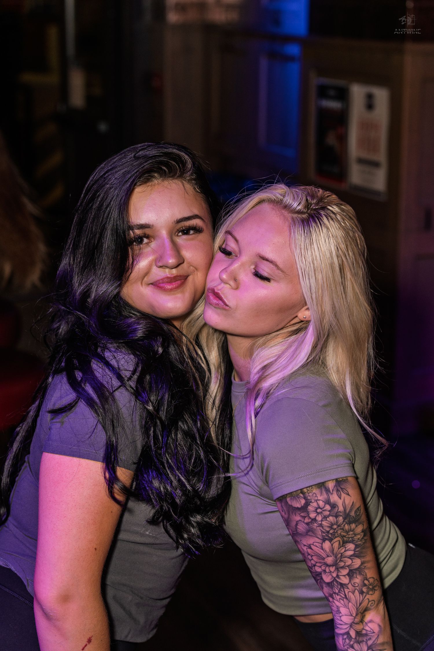 Two women share a playful moment at a nightclub, one with dark hair and one blonde with floral arm tattoo.
