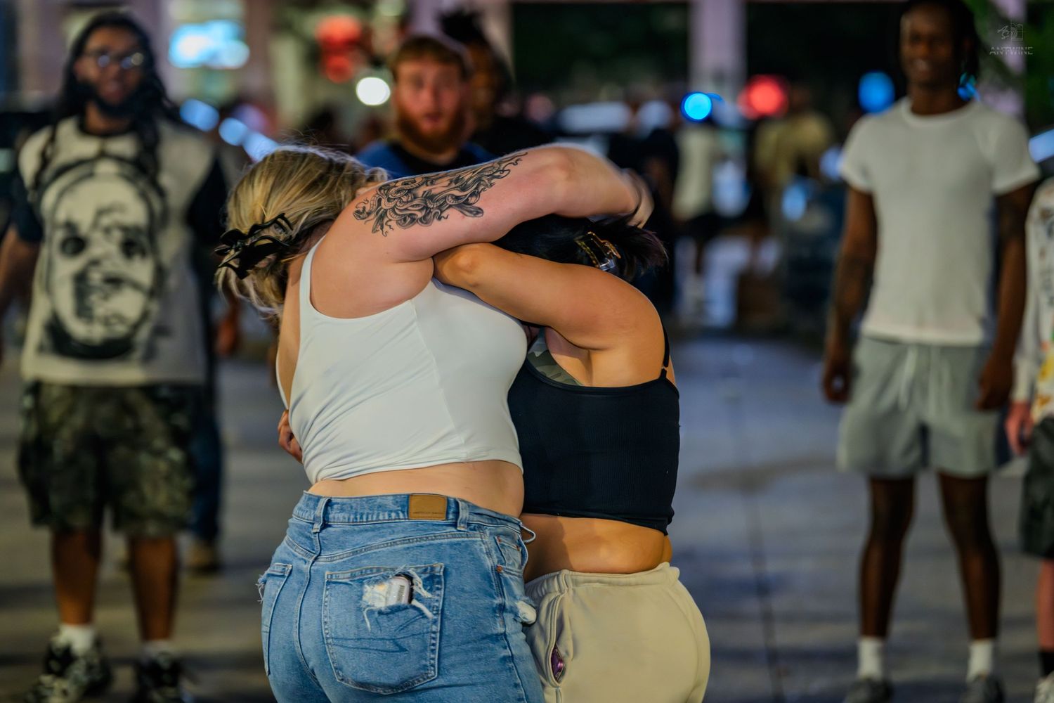 Two women hugging on a busy nighttime street with people gathered in the background.