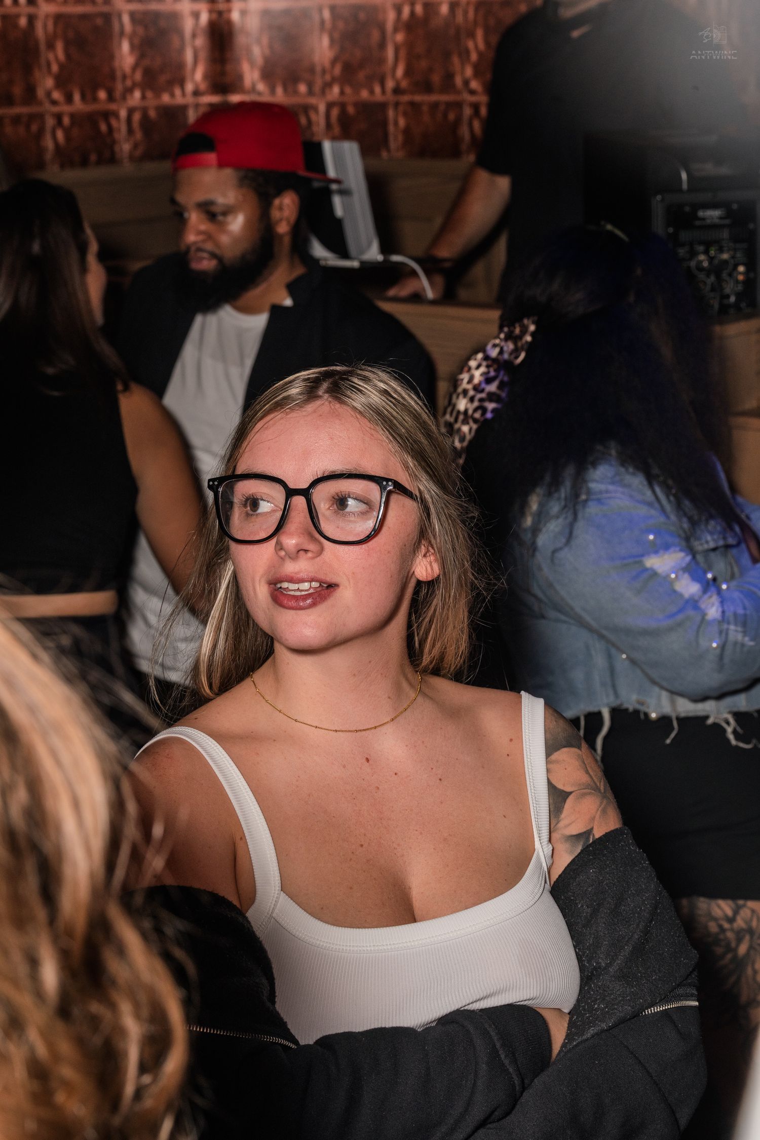Blonde woman with large black glasses smiling at a crowded indoor nightclub party.