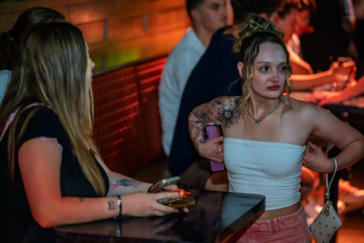 Tattooed woman in white crop top socializes at a dimly lit bar.