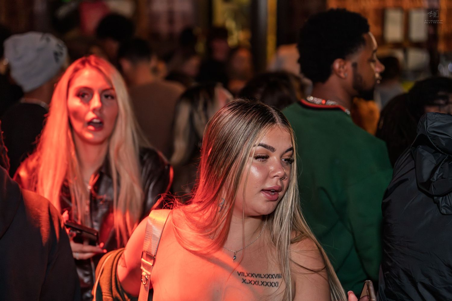 Two blonde women standing in a crowded outdoor nightlife venue illuminated by warm red lighting.