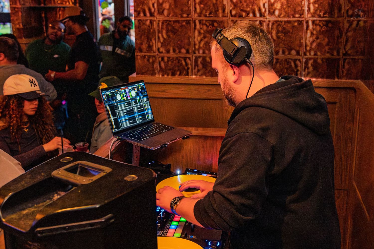 DJ mixing music on equipment with laptop at a crowded St. Patrick's Day bar event.
