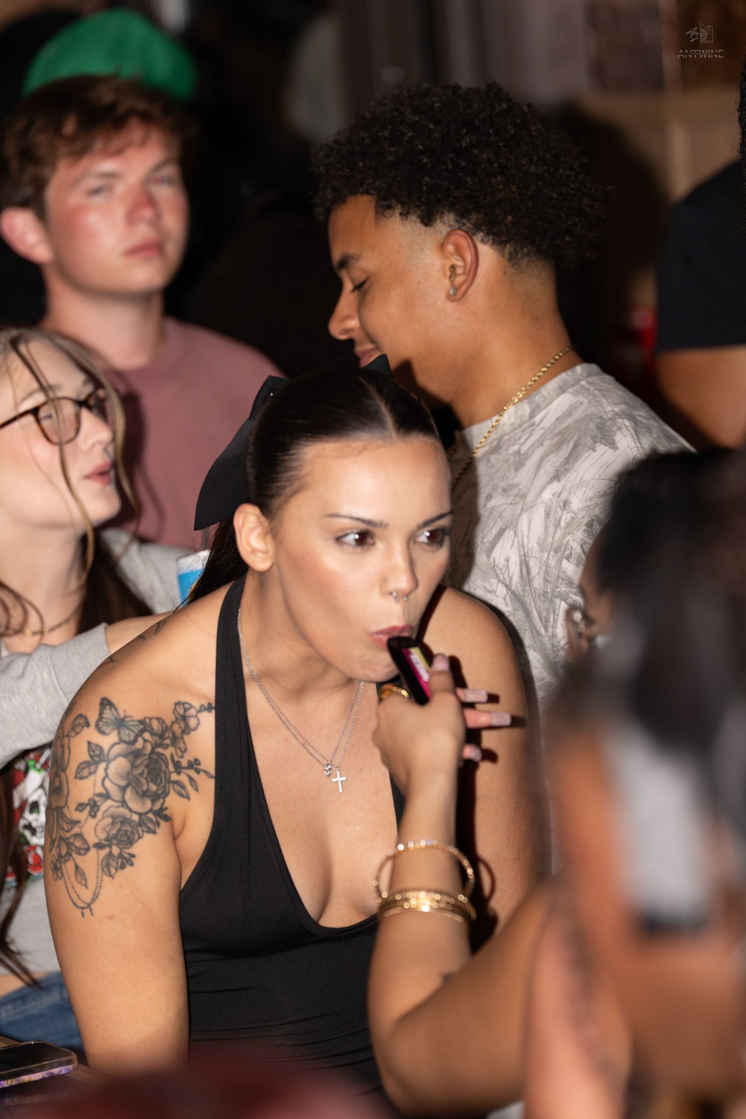 Woman with floral shoulder tattoo drinking at a crowded nightclub bar event.