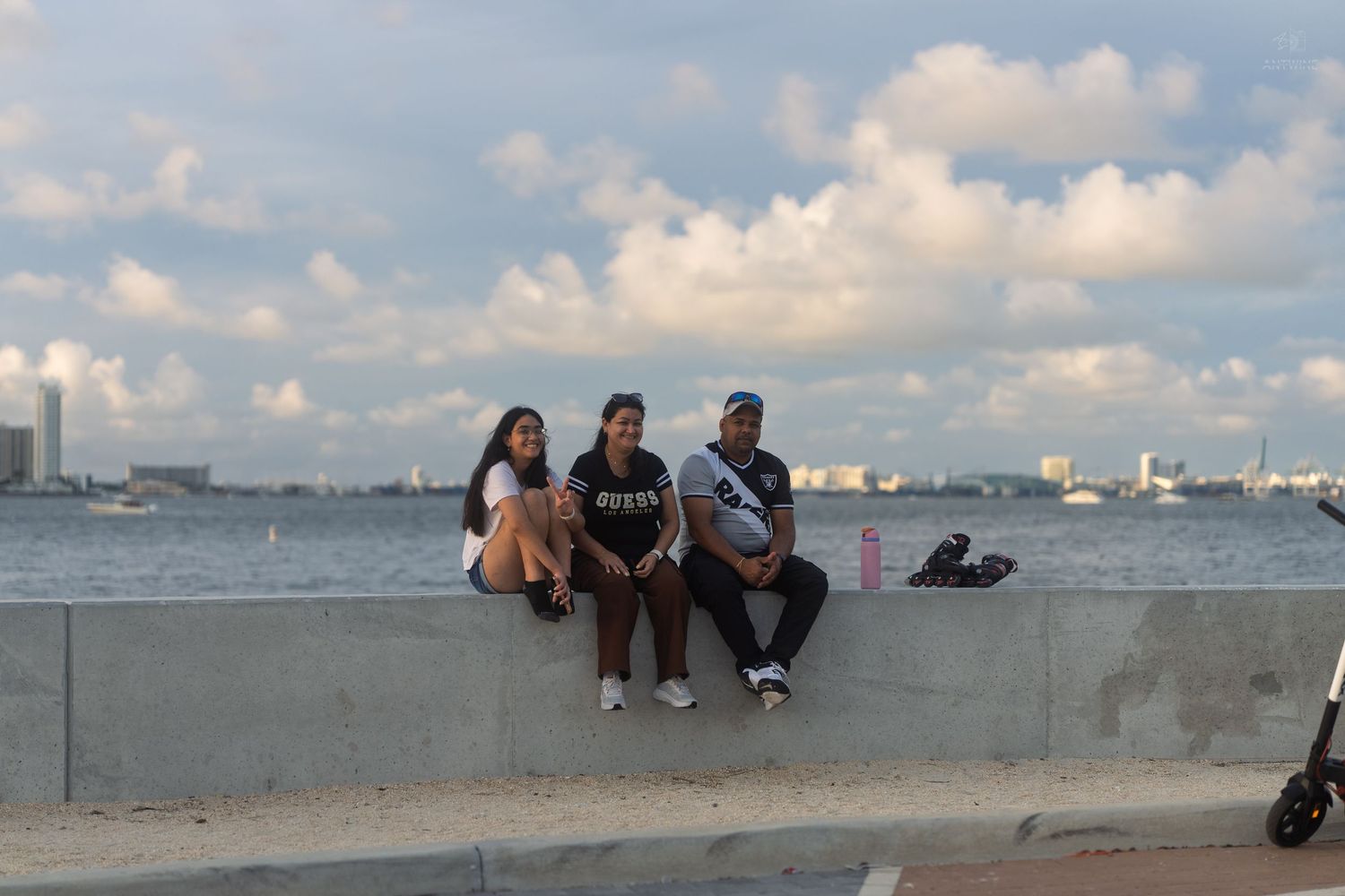 Three friends sitting together on a concrete waterfront wall with scenic bay and cloudy blue sky behind them.
