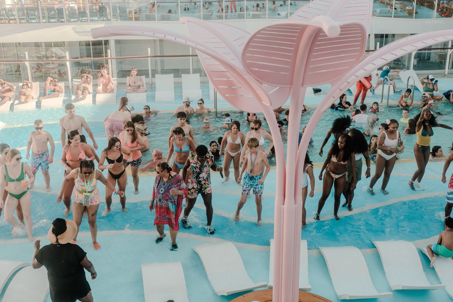 Aerial view of cruise ship pool party with guests dancing near a pink decorative palm tree sculpture.