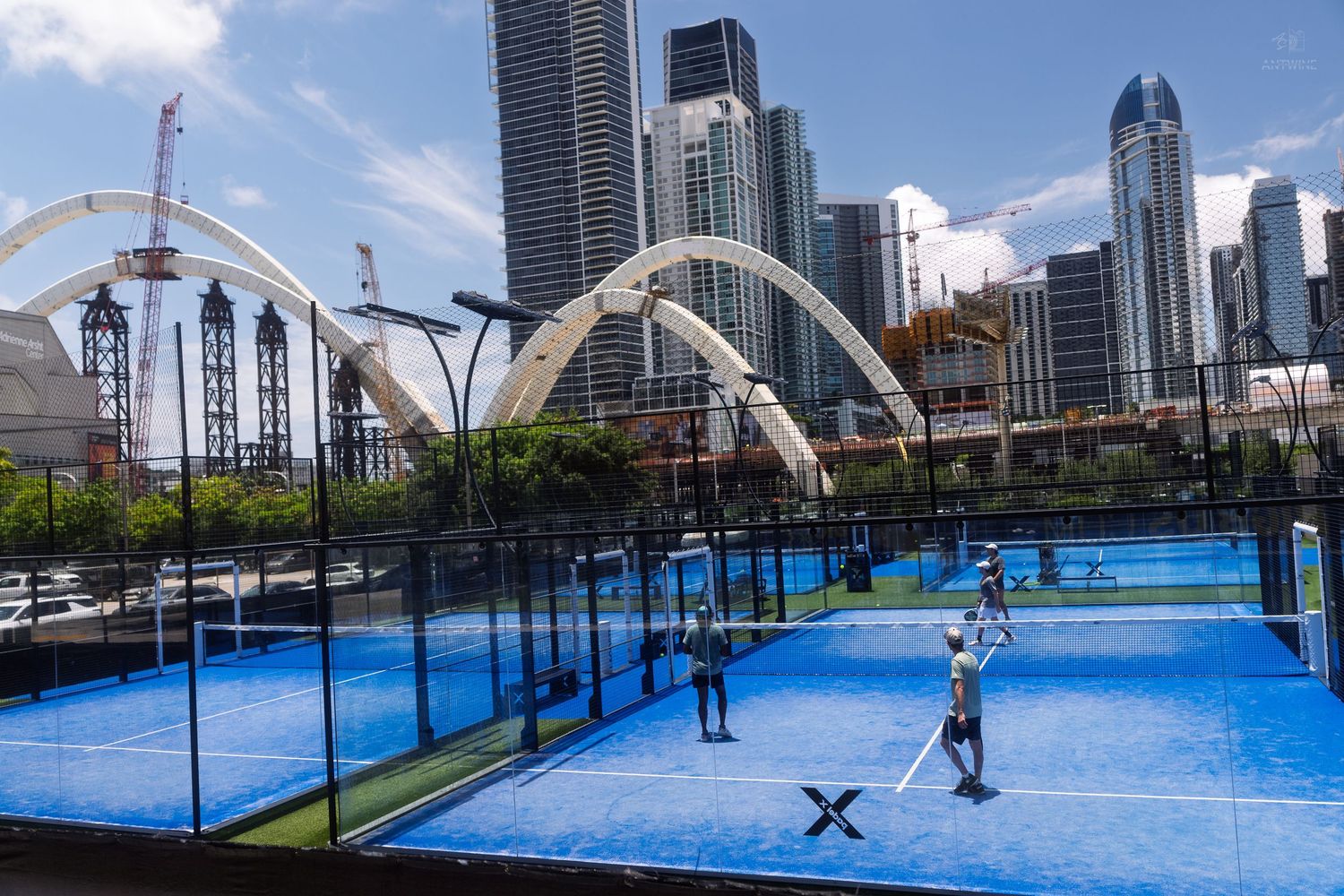 Players compete on blue padel tennis courts with a stunning city skyline and white arch structures in the background.