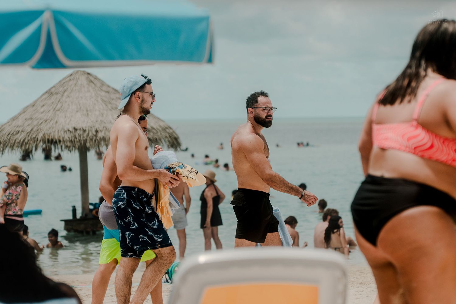 Two shirtless men walking along a crowded tropical beach near a thatched hut.