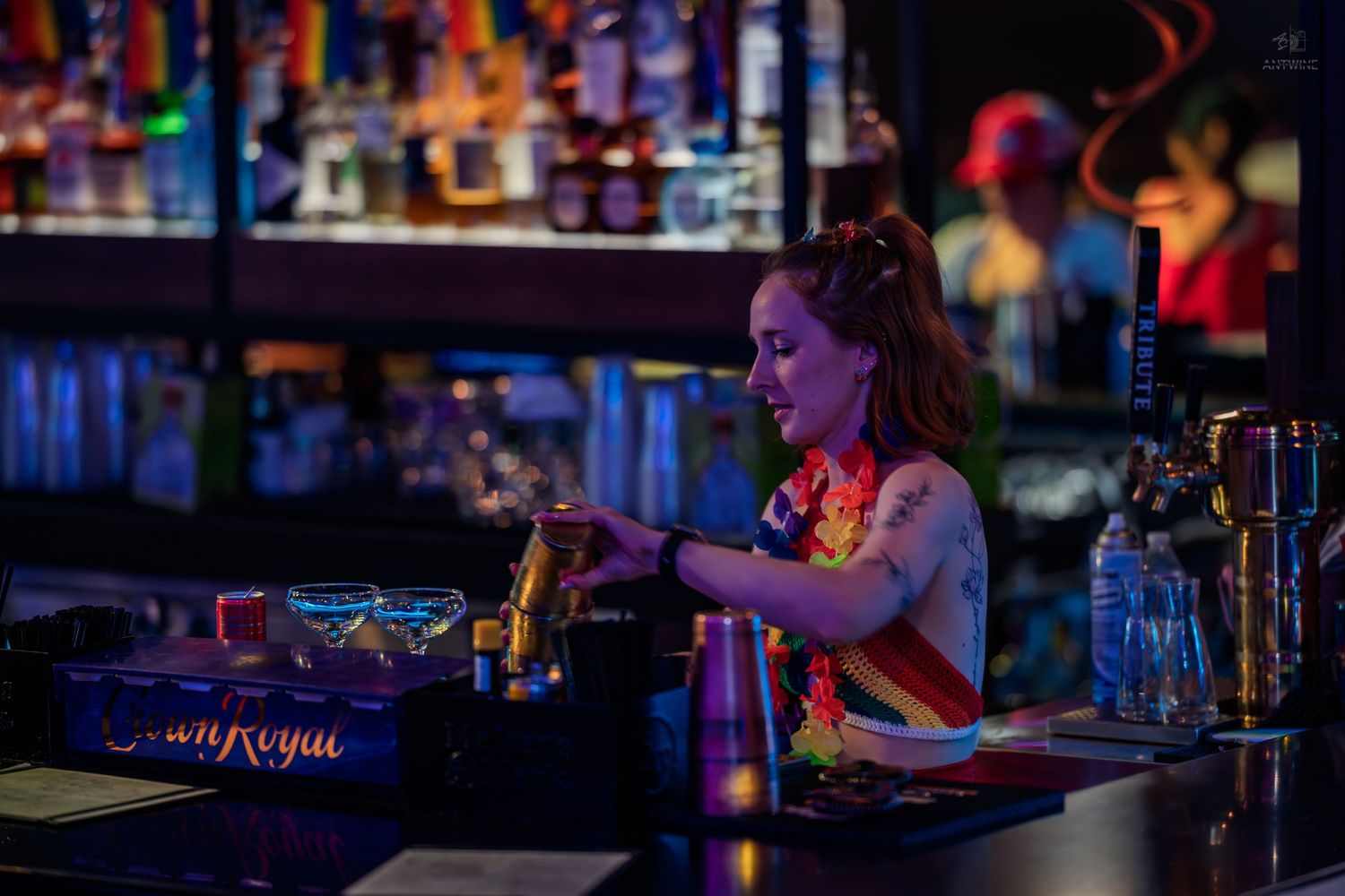 Tattooed female bartender mixing cocktails behind a Crown Royal branded bar under colorful lights.