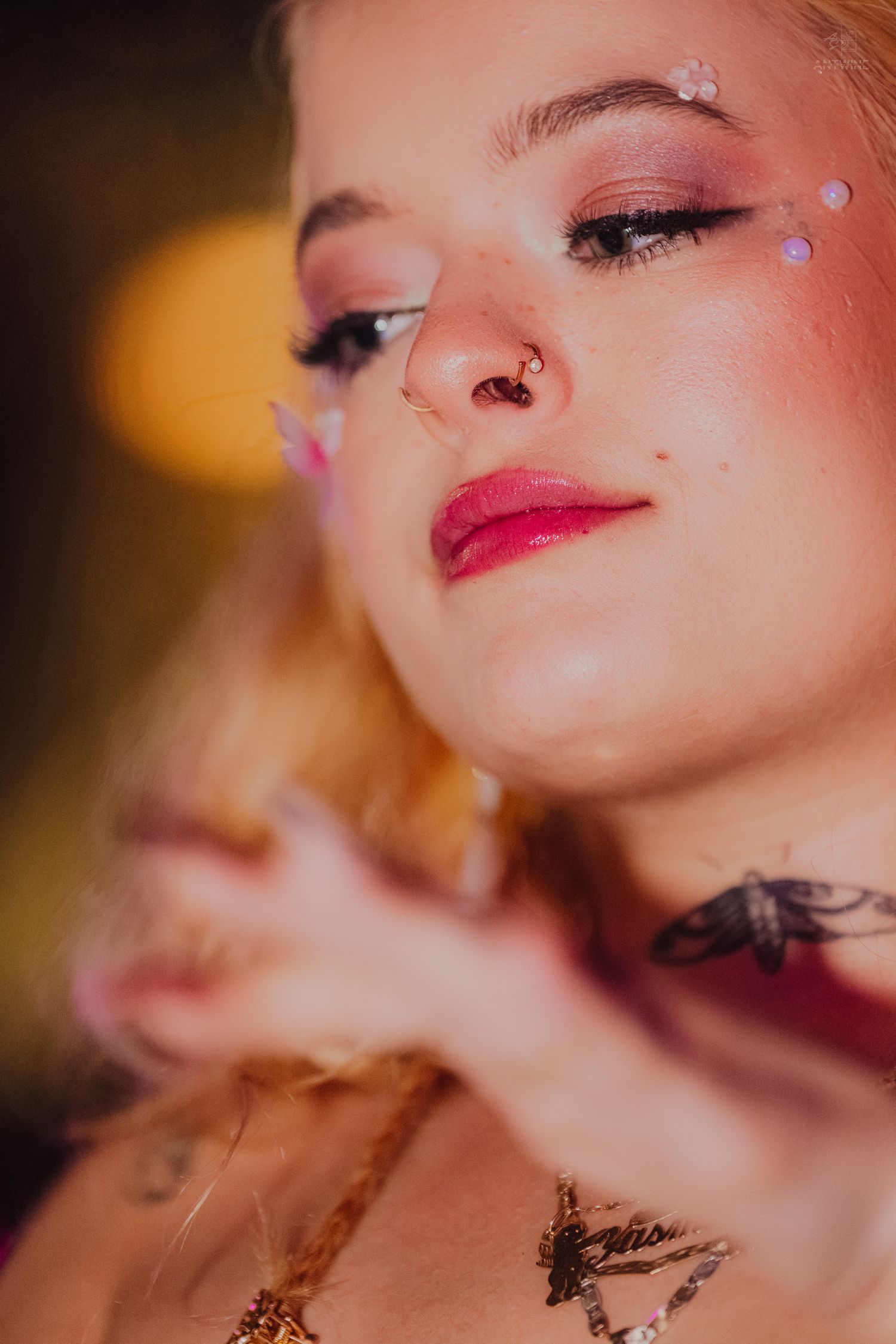 Close-up portrait of blonde woman with glitter face gems, nose ring, red lips, and neck tattoos.