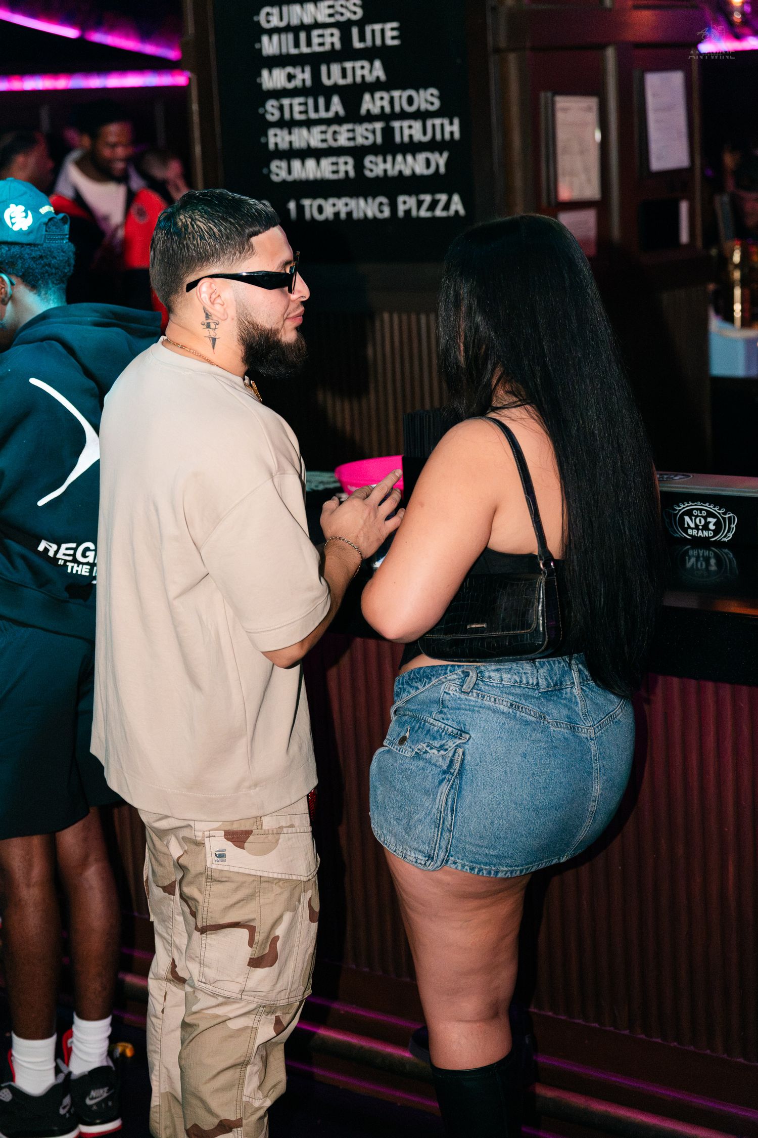Couple talks near a bar menu board listing beers and pizza at a nightclub.