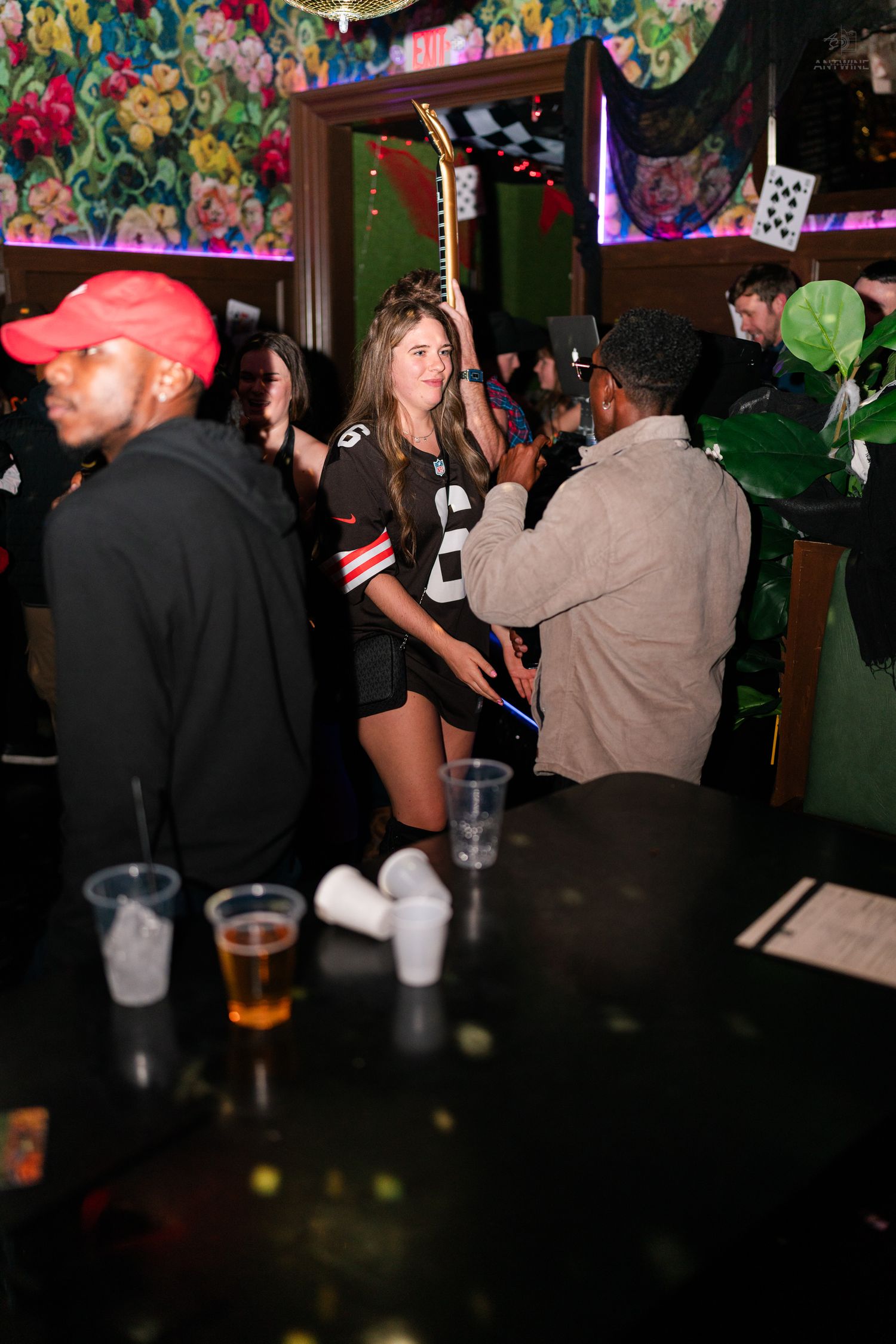 Woman in football jersey costume socializing at a crowded nightclub bar with colorful decor.