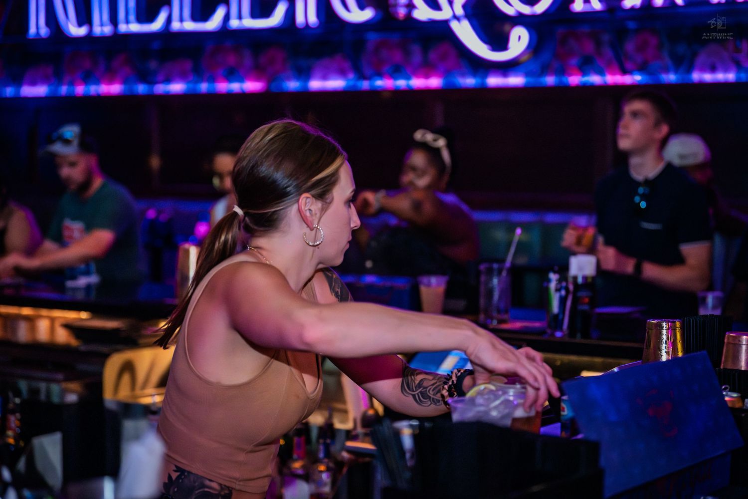 Female bartender pouring drinks at a neon-lit bar with signage in background.