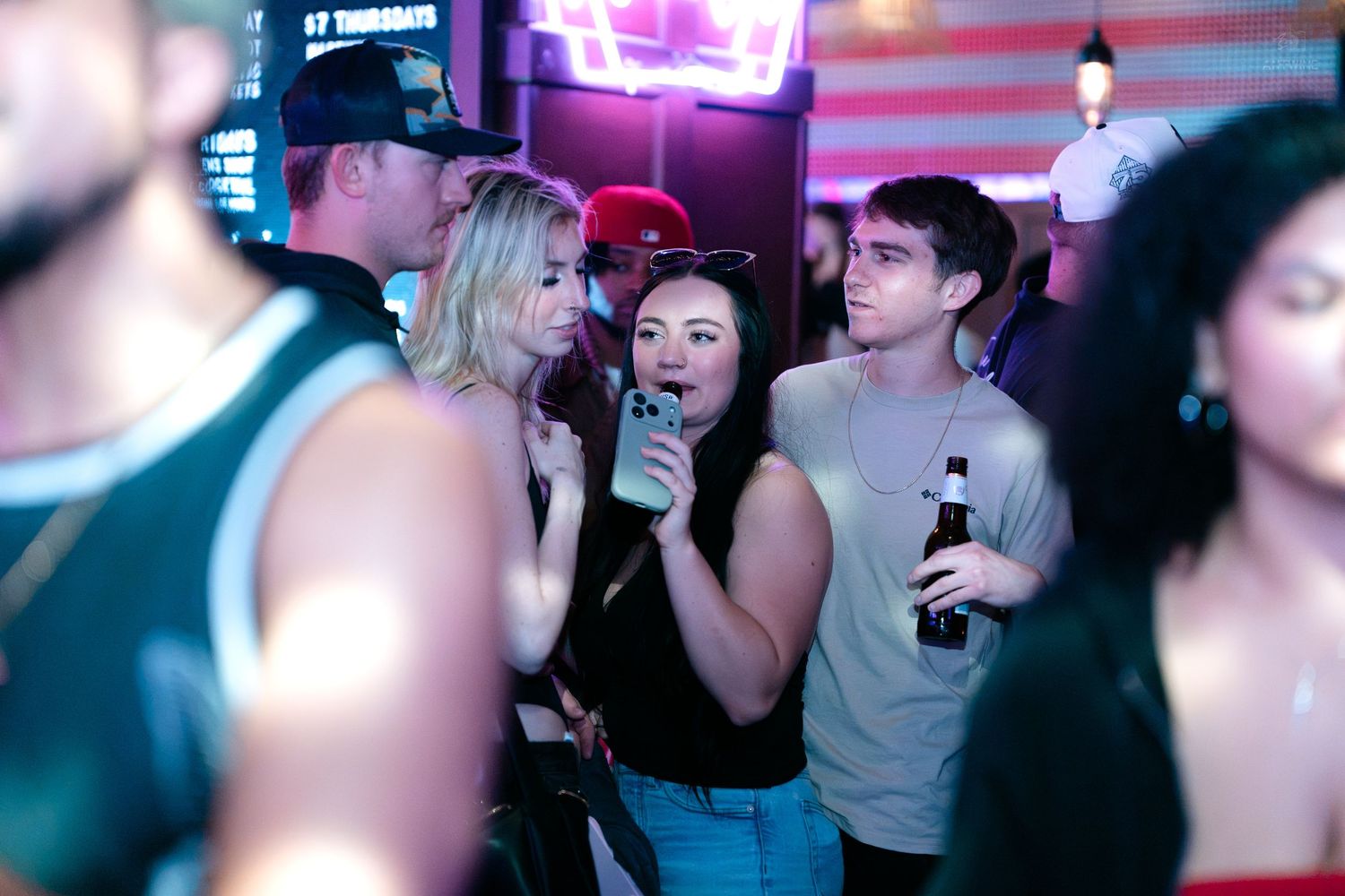 Group of young adults socializing and drinking at a neon-lit nightclub bar.