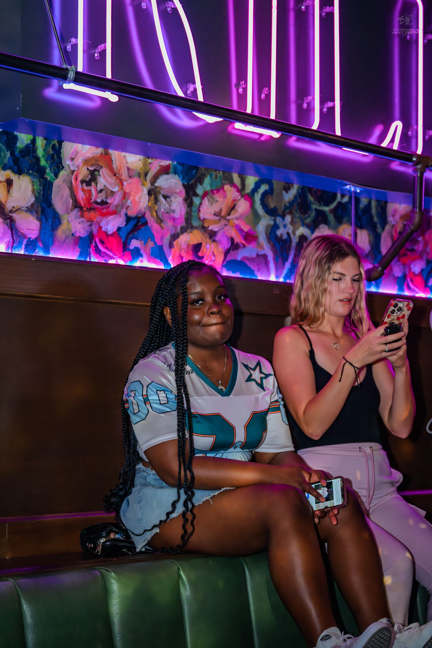 Two women sitting in a green booth at a nightclub under pink neon signs and floral decor.