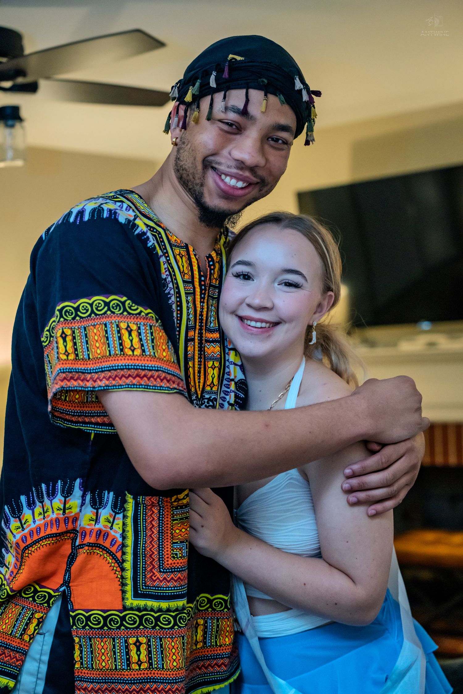 A young man in a colorful African dashiki shirt smiles while hugging a happy young girl indoors.