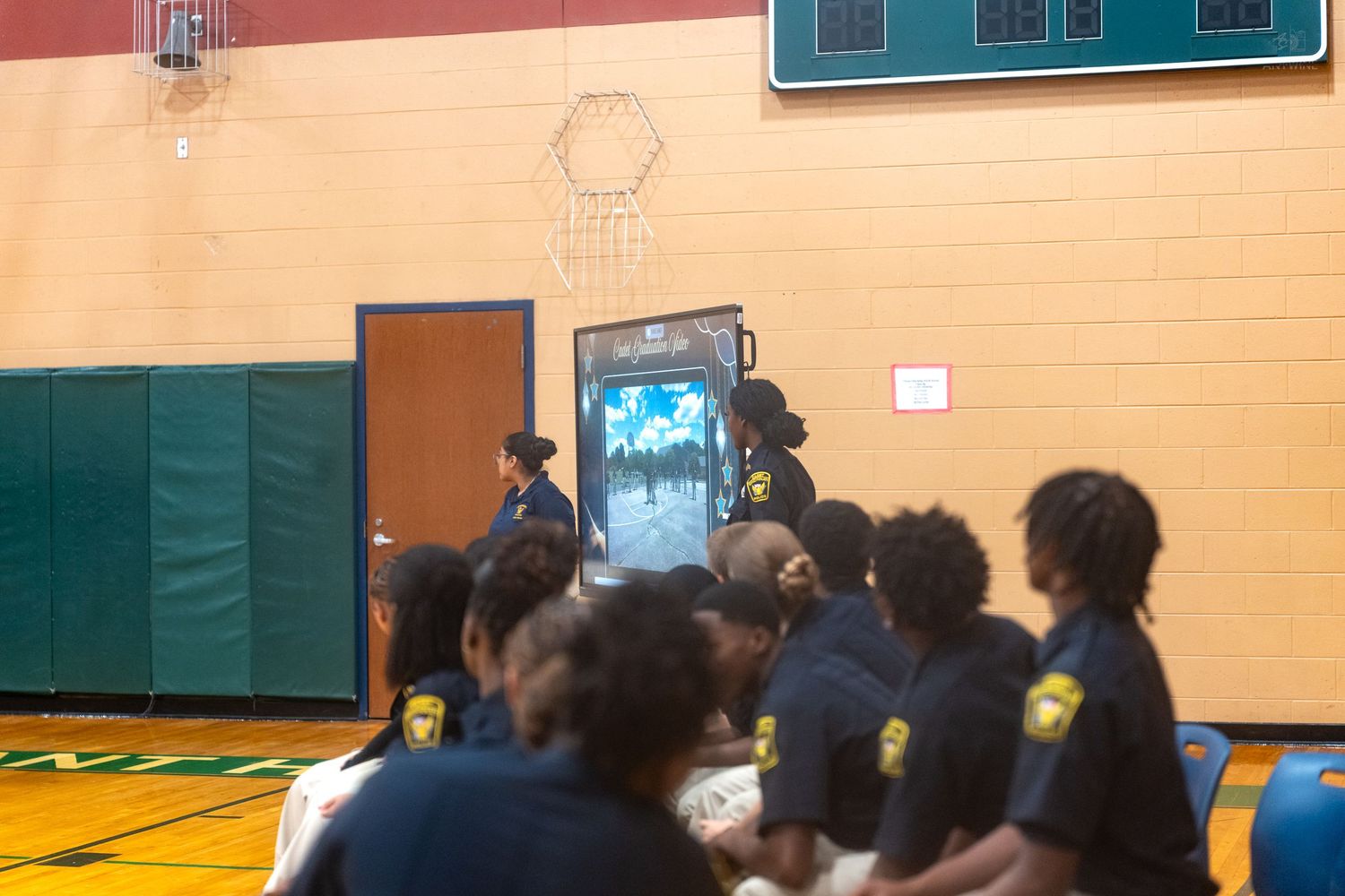 Students in uniforms gather in a school gymnasium watching a presentation on a large screen display.