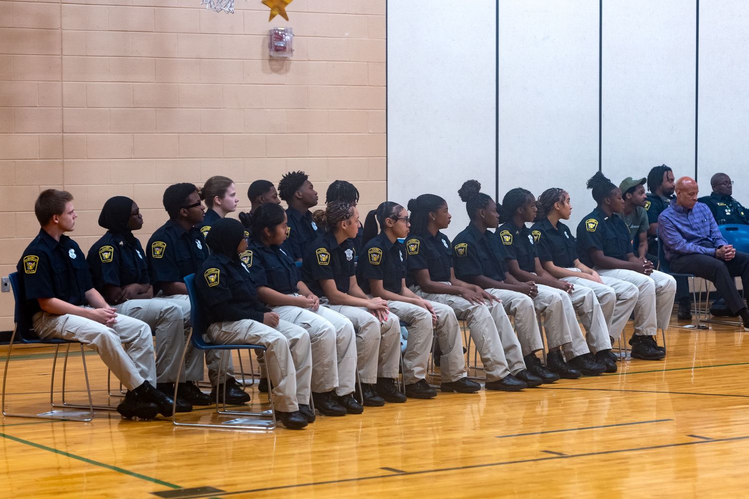 Young students in matching navy uniforms sit in rows on a gymnasium floor during a formal school ceremony or graduation.