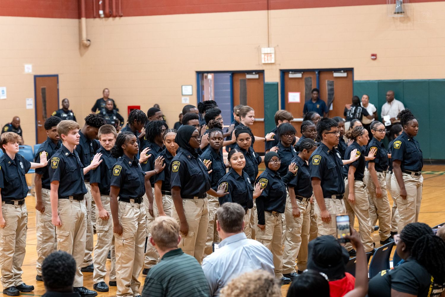 Young JROTC cadets in matching uniforms recite an oath during an indoor school ceremony in a gymnasium.