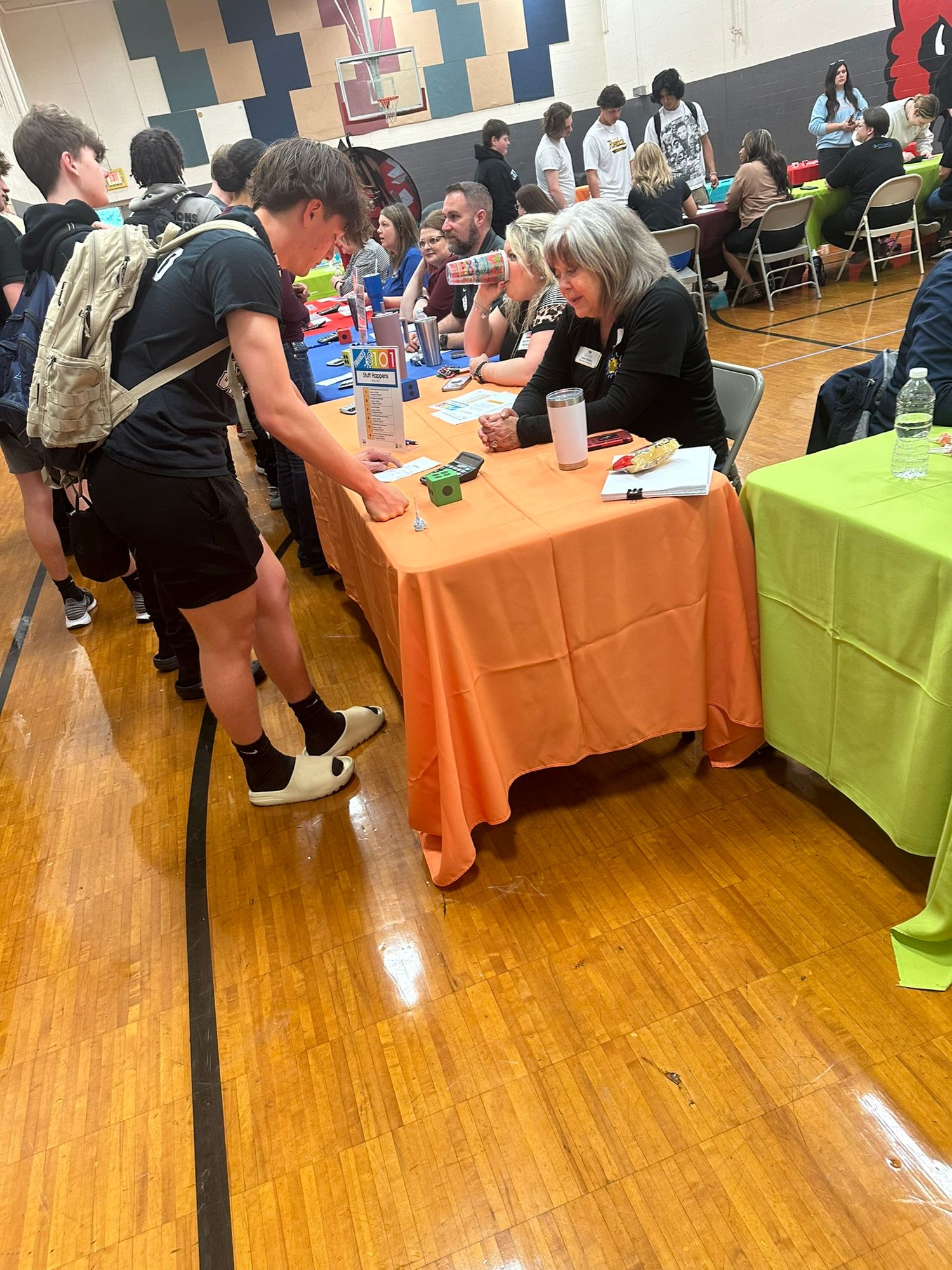 Students browse booths at a school career fair with orange and green tables in a gym.