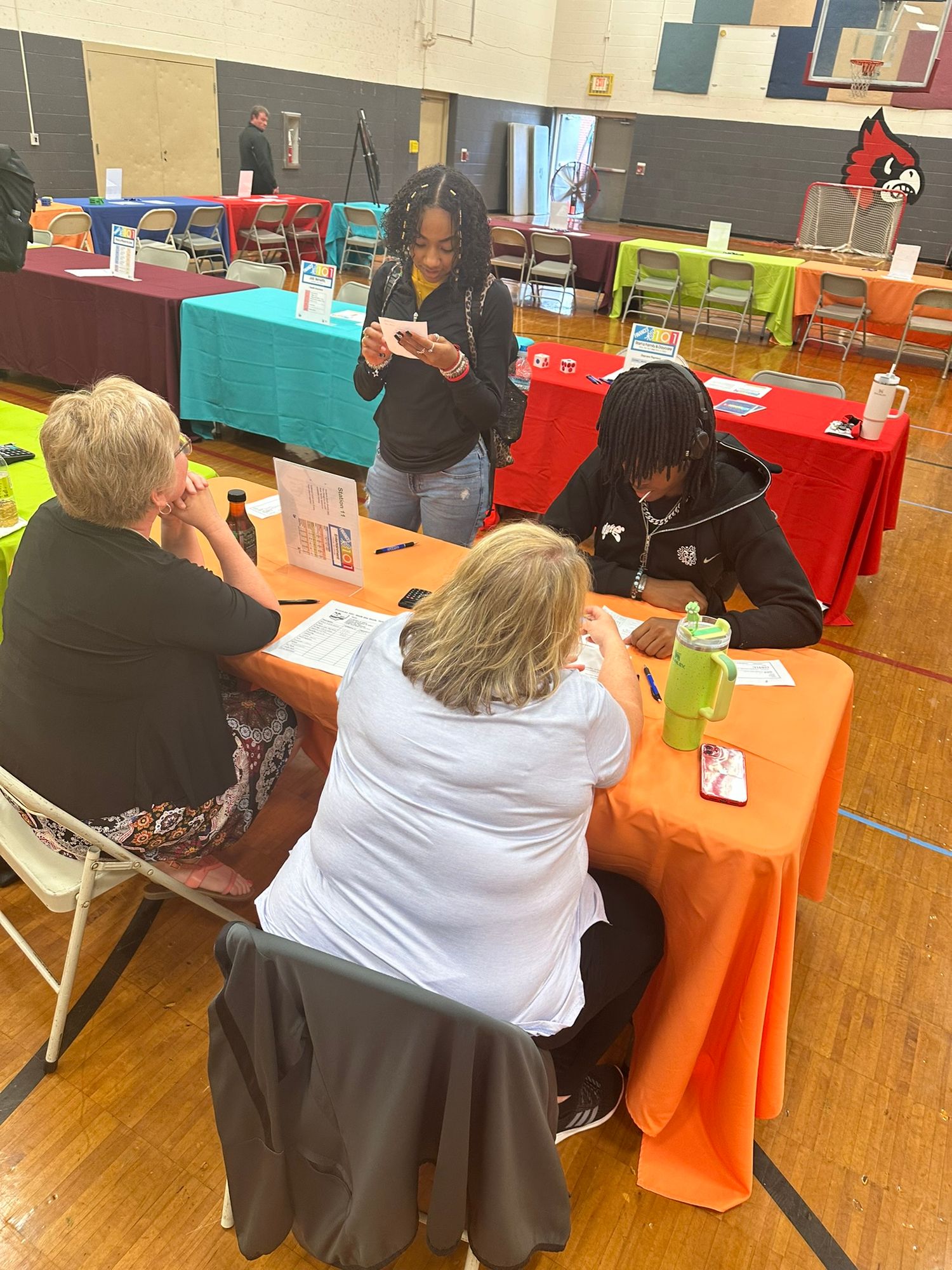 People gathered around orange-clothed table in school gymnasium during a community event or job fair.
