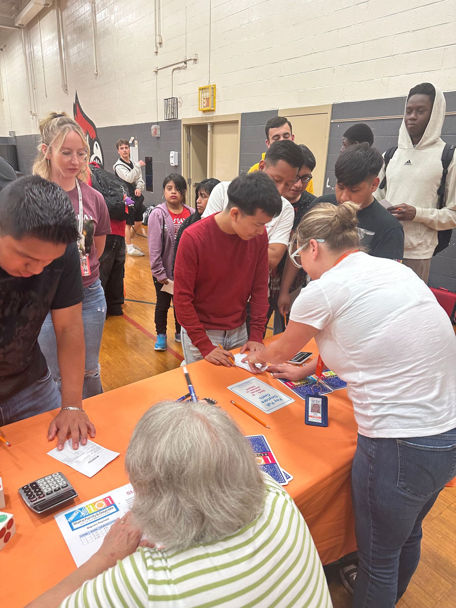 Students and adults gather around orange tables in a gymnasium, signing papers at what appears to be a school registration event.