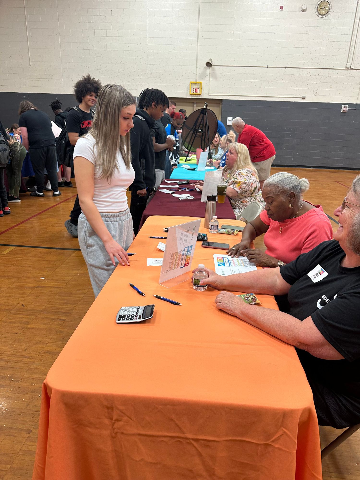 Students engage with educators at an orange table during a school fair event in a gymnasium.