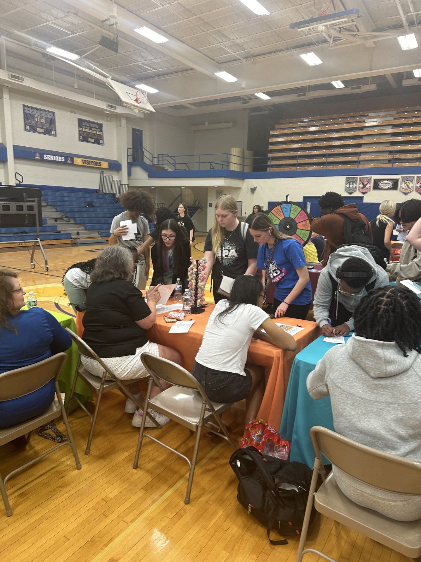 Students gather around tables in a school gymnasium during an educational fair event with a colorful prize wheel.