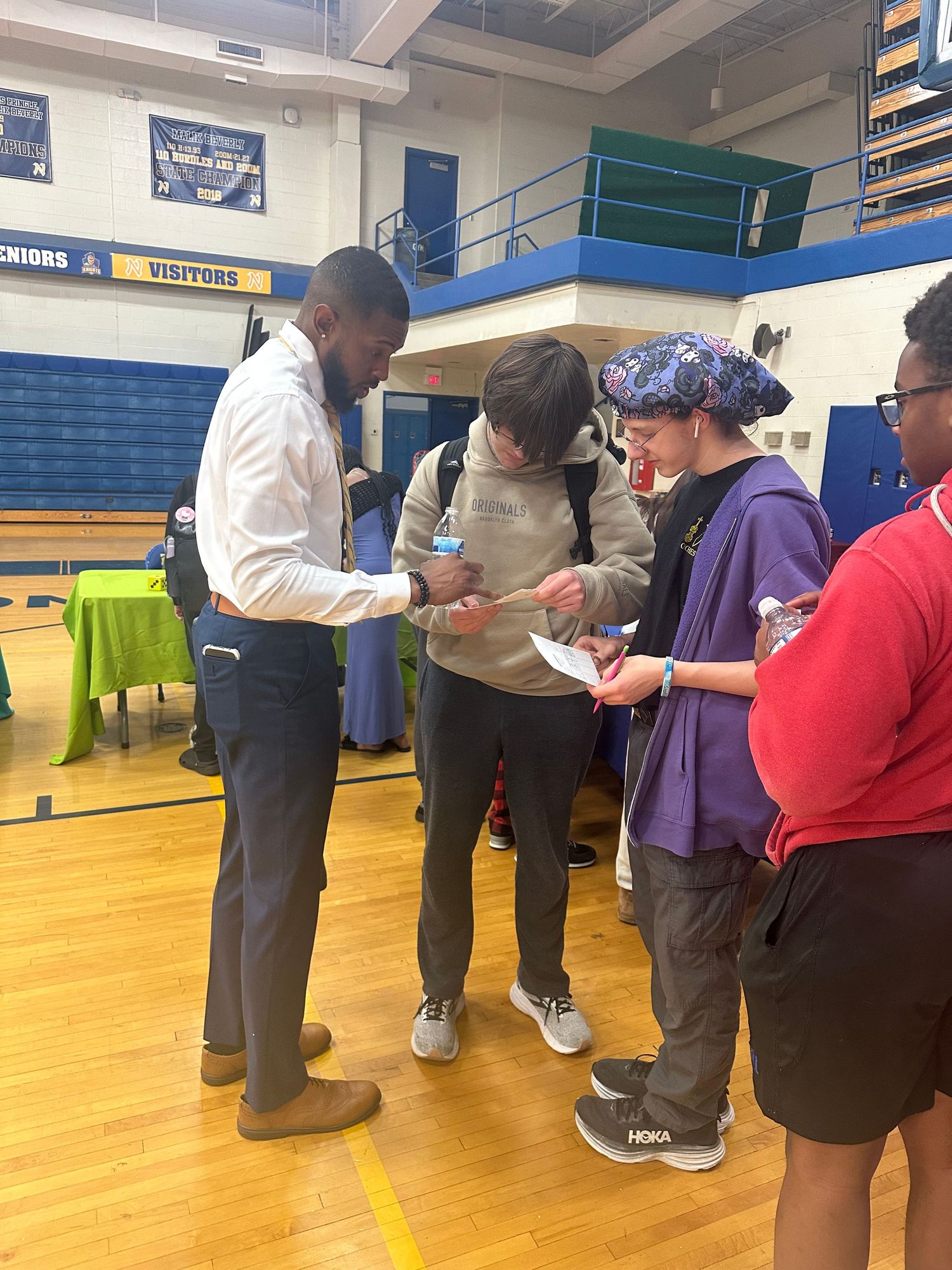 A man in a white shirt reviews papers with students wearing backpacks inside a school gymnasium event.