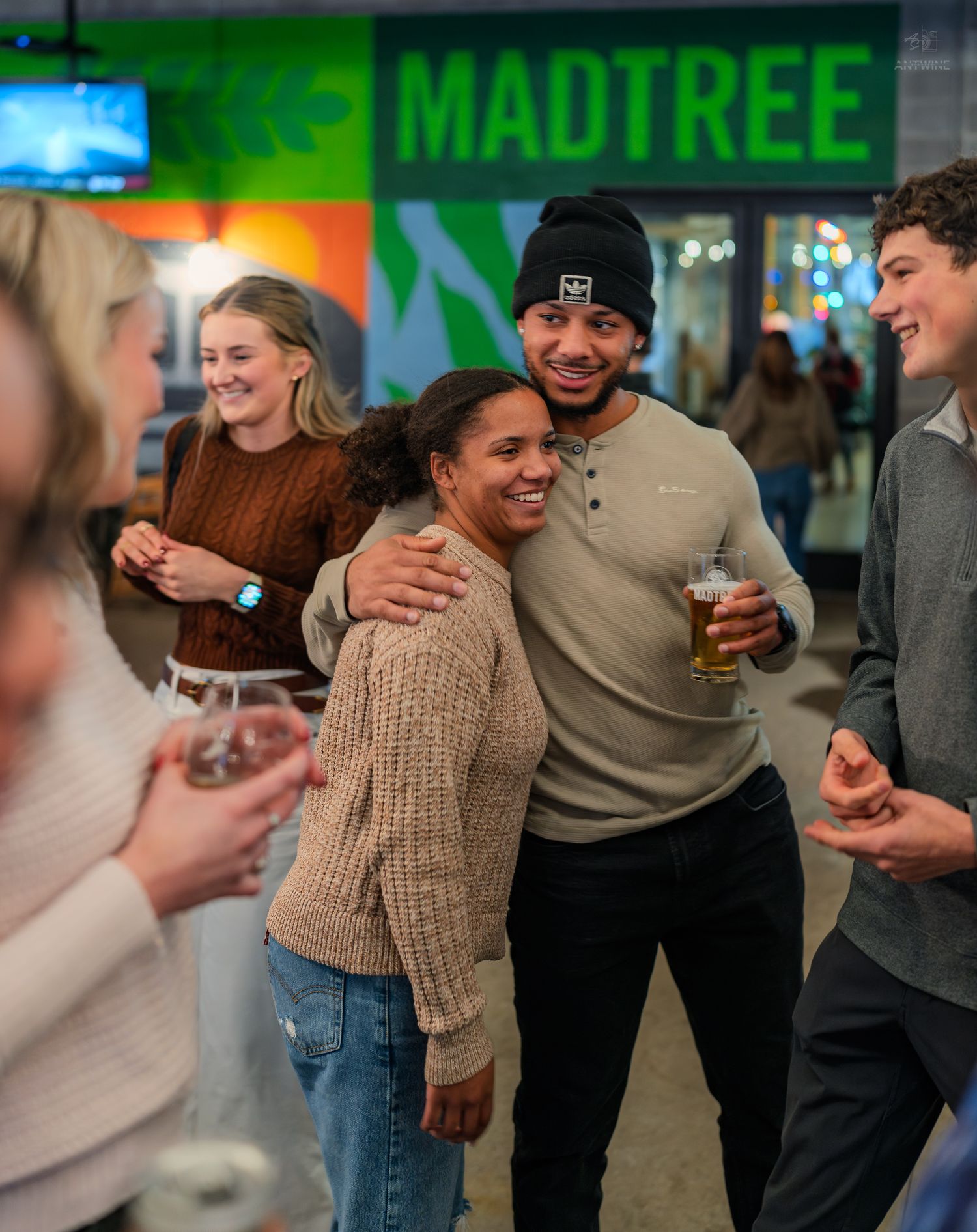 A couple embraces and smiles at a MadTree Brewing social event while friends laugh around them.