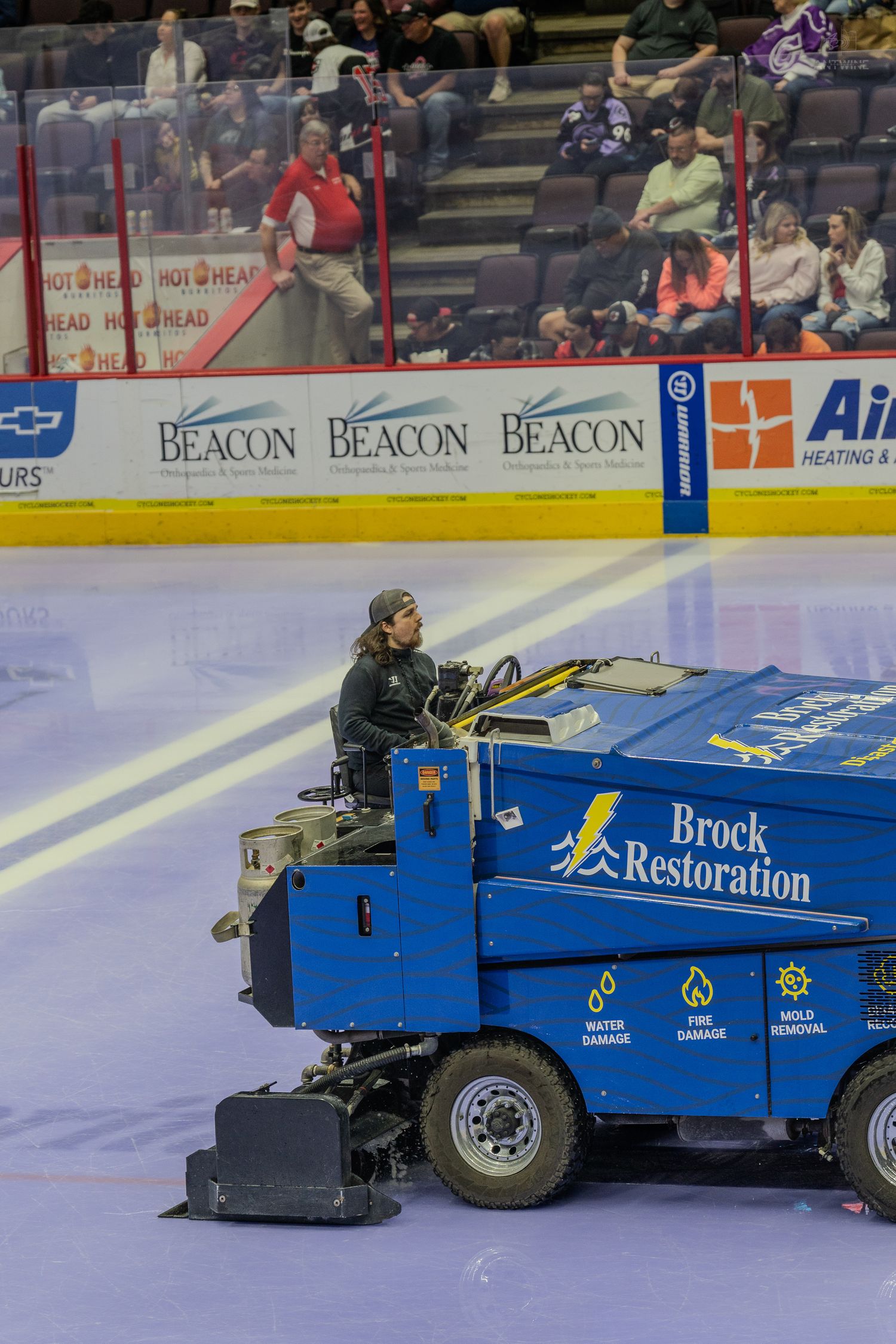 A driver operates a blue Brock Restoration Zamboni on the ice at a hockey arena near Beacon advertisement boards.