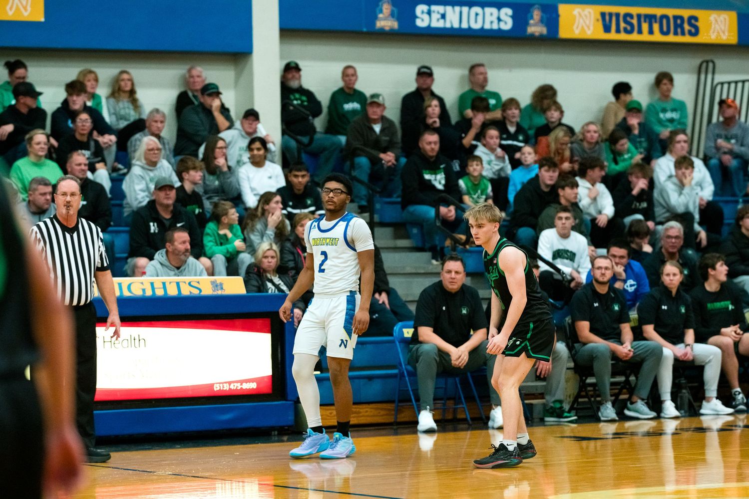 High school basketball player number 2 in white faces green-uniformed opponent near the sideline with packed stands.