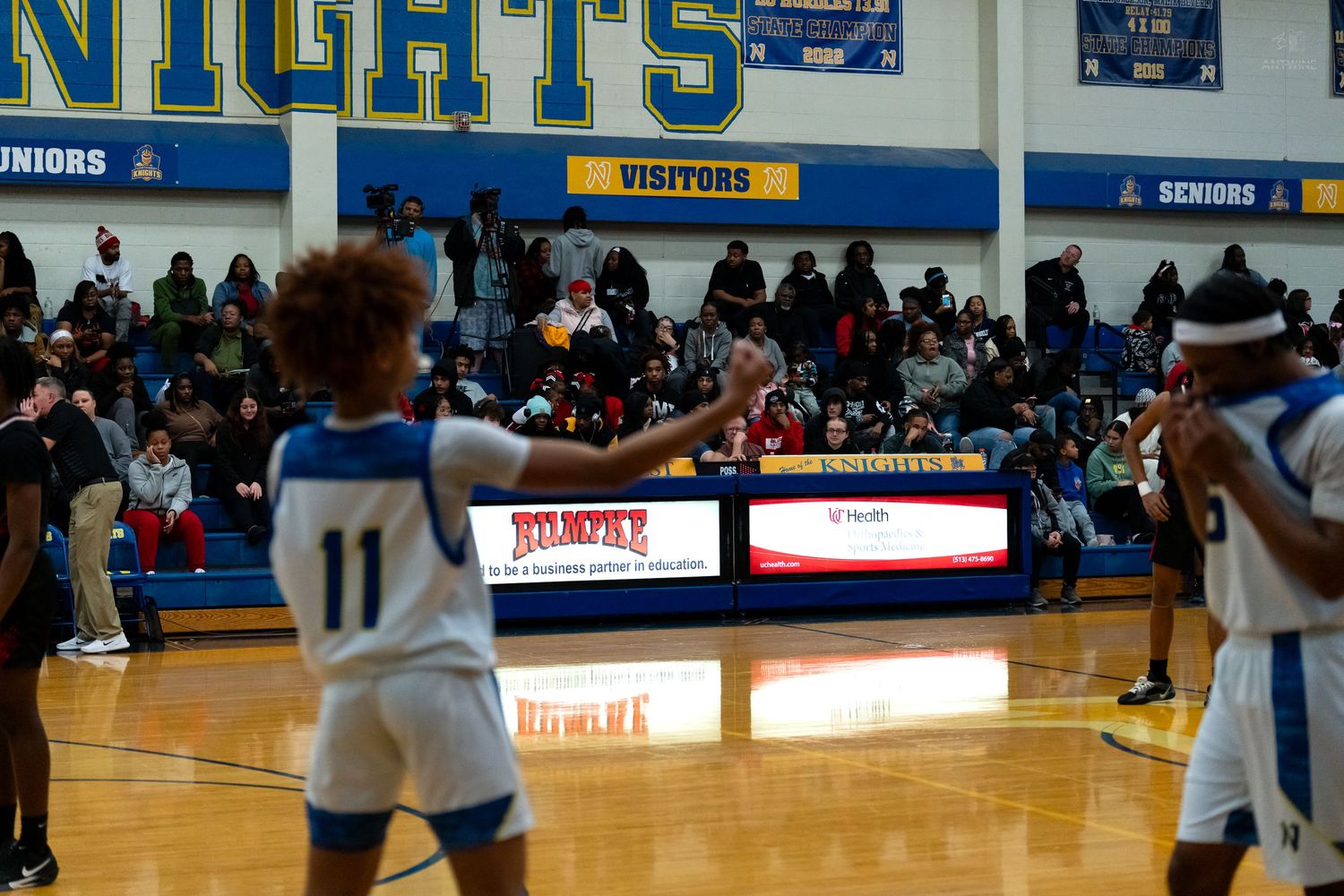 High school basketball game in a Knights gymnasium with players on court and packed spectator stands.
