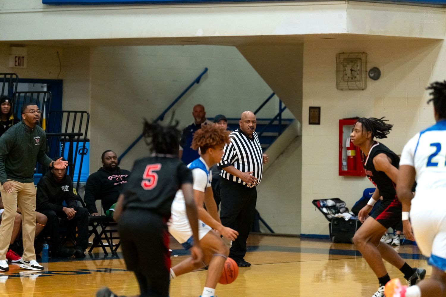 Basketball referee watches players during a high school game with player number 5 in red uniform dribbling.