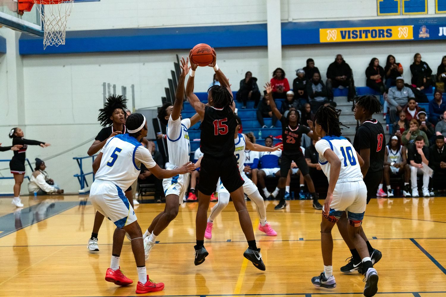 Red-uniformed player leaps for basket surrounded by blue team defenders during intense high school basketball game.