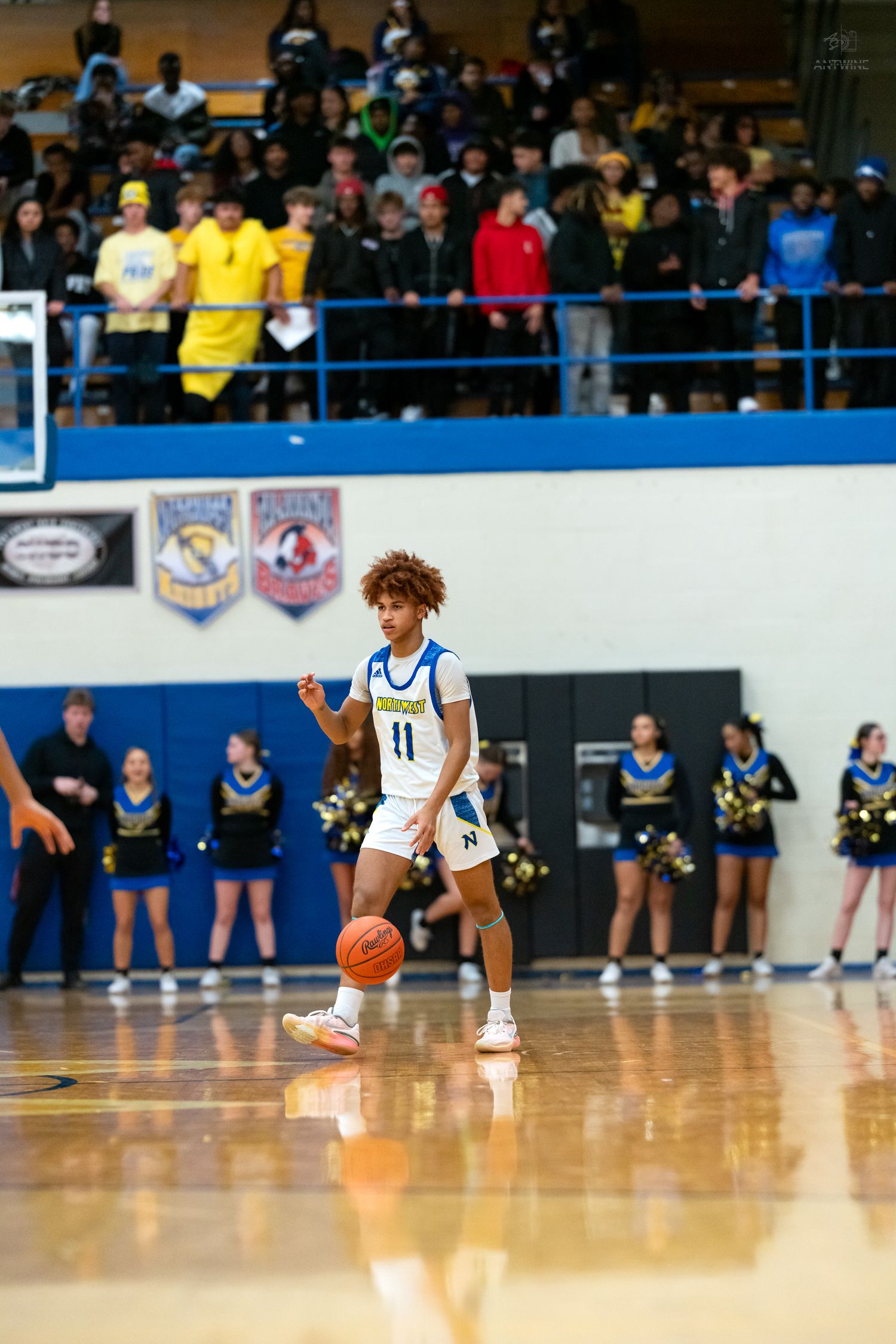 Player #11 in white and blue uniform dribbles basketball during high school game with packed crowd.
