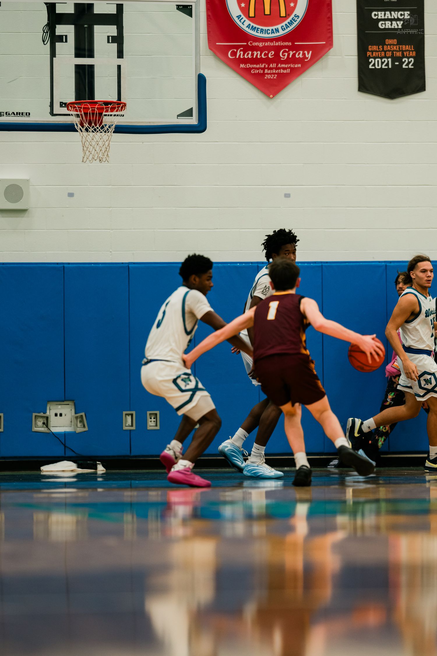 High school basketball players compete on a blue court as a defender guards an opponent dribbling the ball.