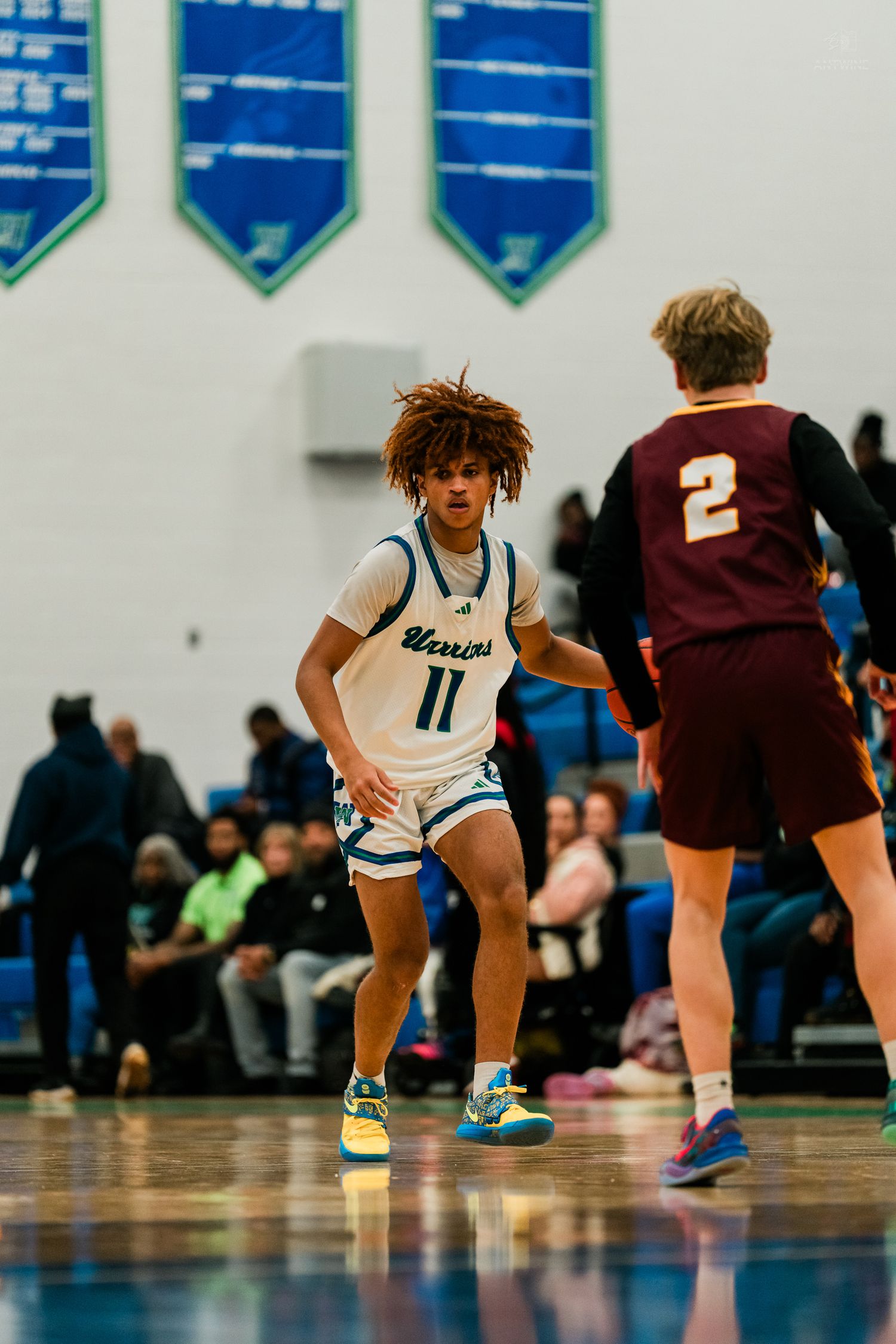 Warriors player number 11 in teal uniform plays defense against maroon-uniformed opponent number 2 in gymnasium.