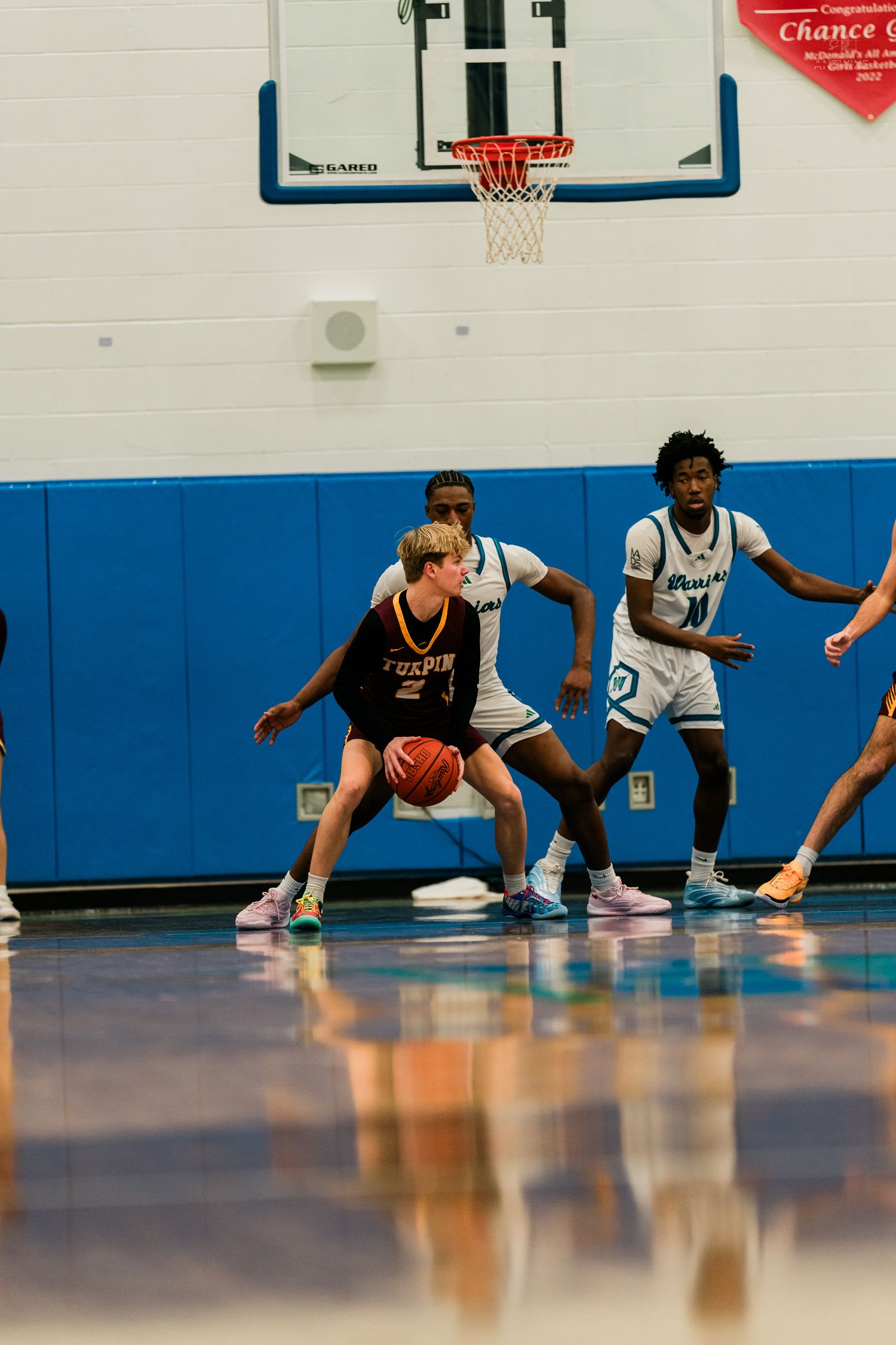 A Turpin player in maroon dribbles while defended by two players in white uniforms near the basketball hoop.