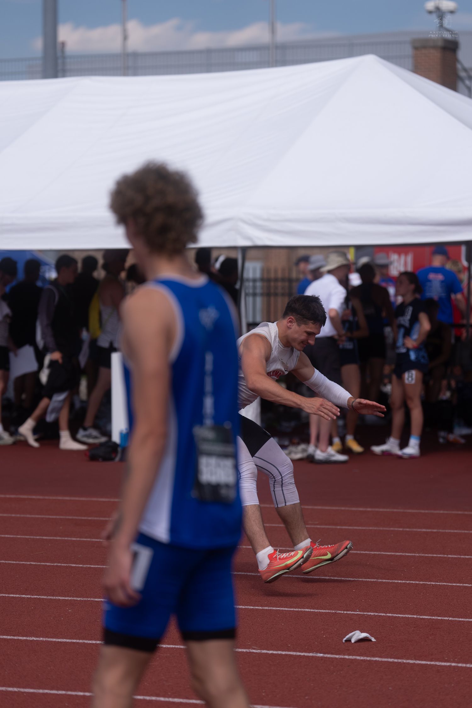 Male athlete in white uniform performs a triple jump approach on a red track at a field competition.