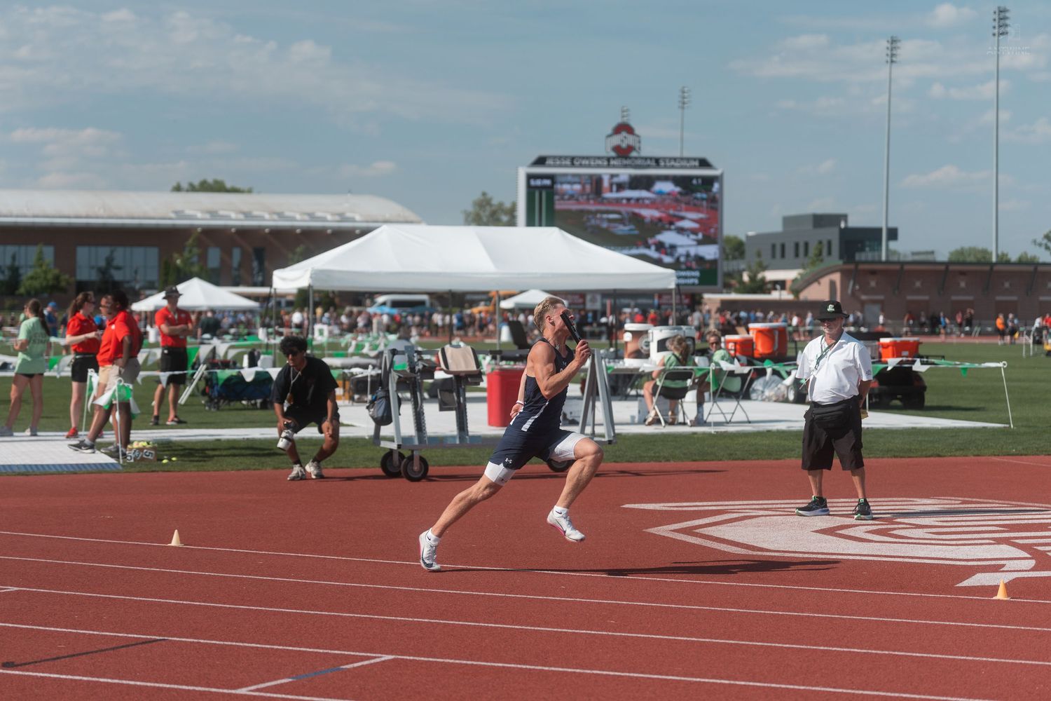 Male runner in navy uniform sprints down red track lane during outdoor track and field competition.