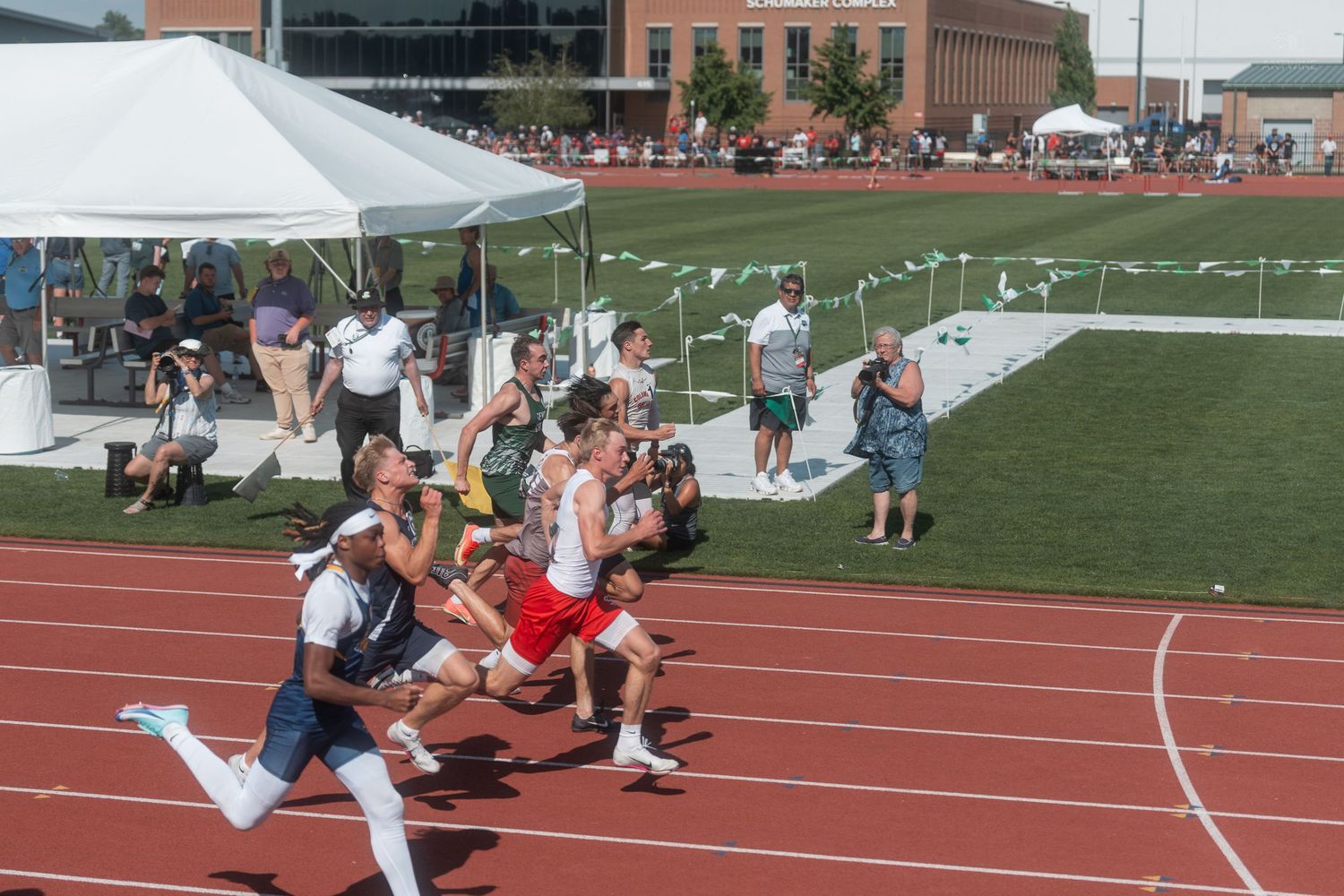 Runners sprint around a red track during a competitive outdoor track and field meet with spectators watching.