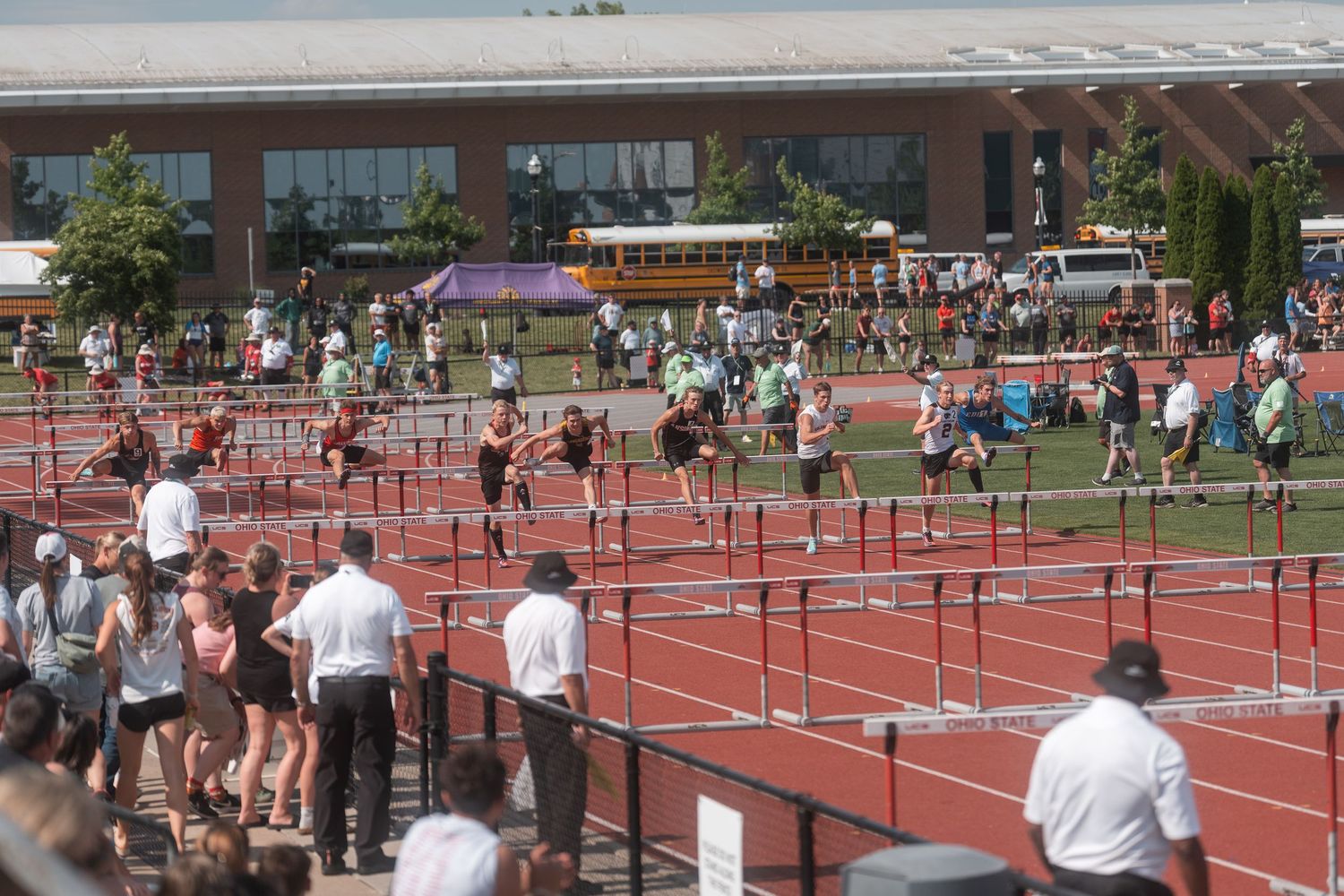 Competitors clear hurdles on red track at outdoor high school track meet with yellow school bus in background.