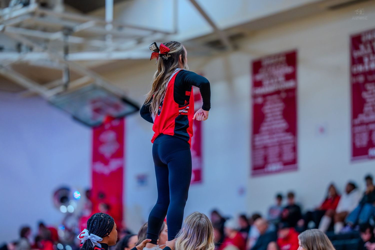High school cheerleader performs an elevated stunt in black and red uniform during a basketball game.